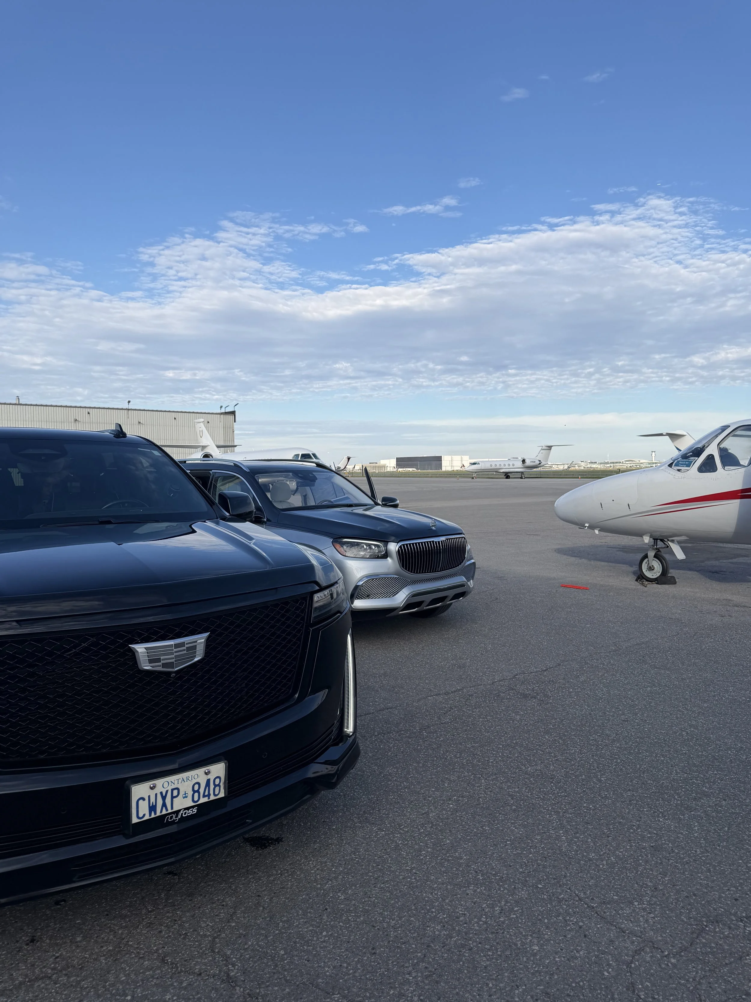 A lineup of vehicles at an airport, including a black Cadillac, a gray car, and a white private jet with a red stripe parked on the tarmac under a partly cloudy sky.