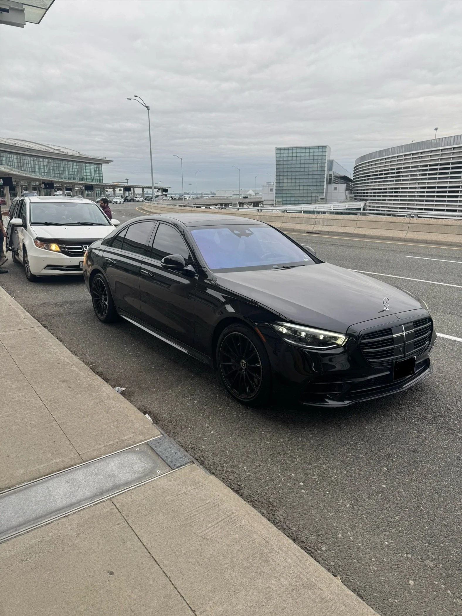Black Mercedes sedan parked on the street near the sidewalk, with a white Honda vehicle and two people standing nearby. In the background, there are modern buildings, a raised highway, and cloudy sky.