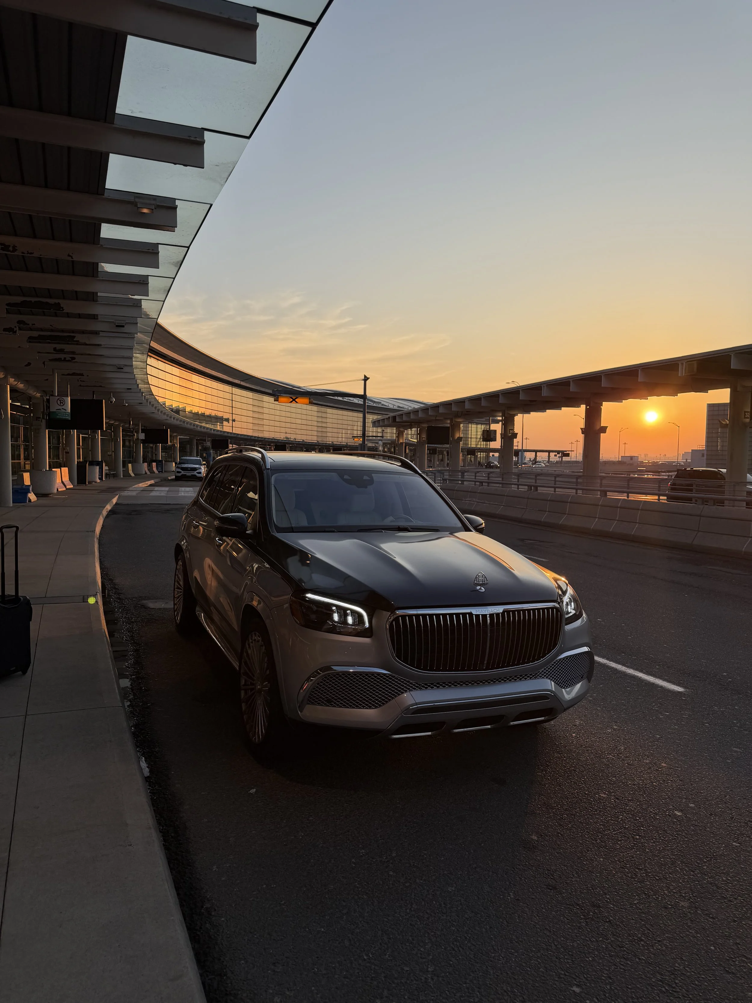 A parking lot at sunset with a black luxury car parked near a sidewalk and a modern building with a glass facade reflecting the sunset.