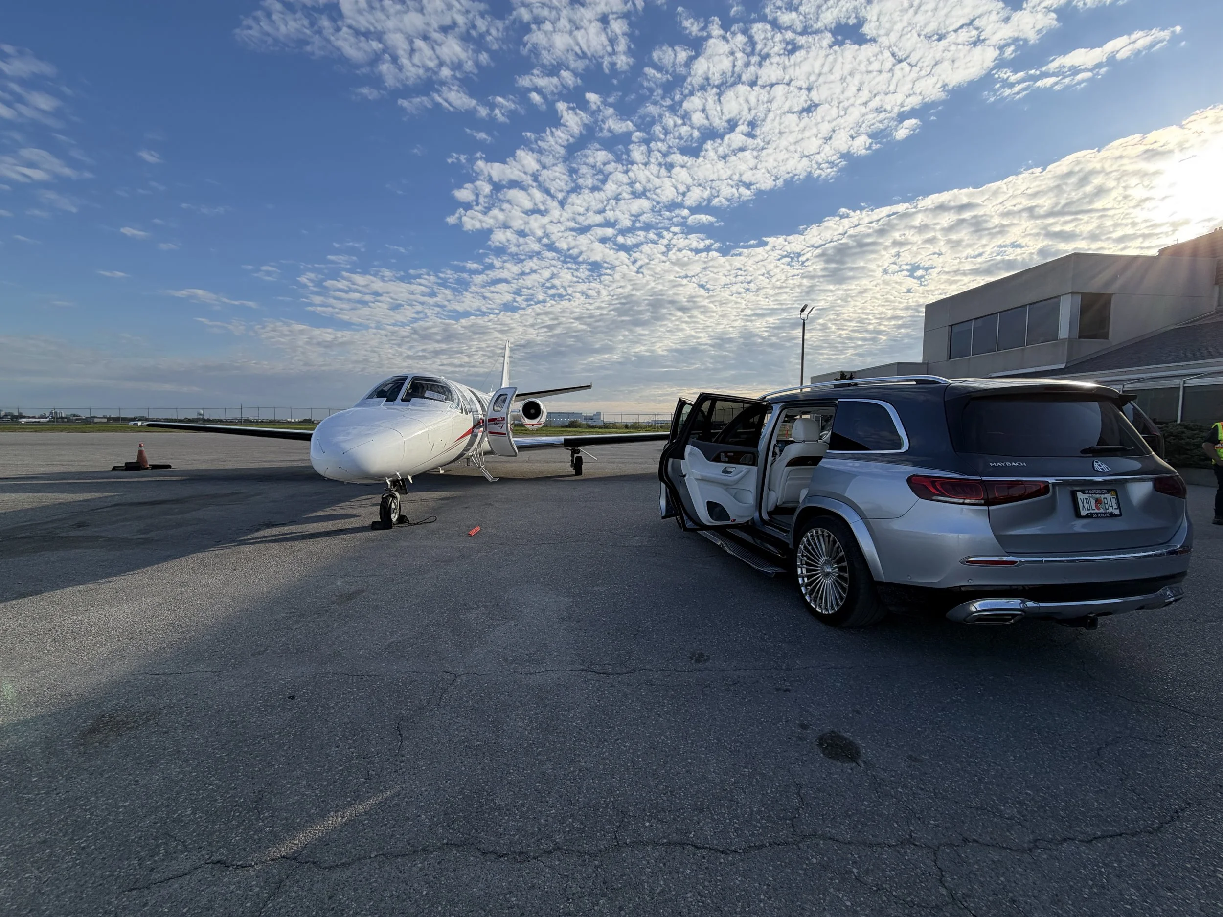 A small private jet parked on an airport tarmac next to a modern building, with a car open door nearby. The sky is partly cloudy with the sun shining.
