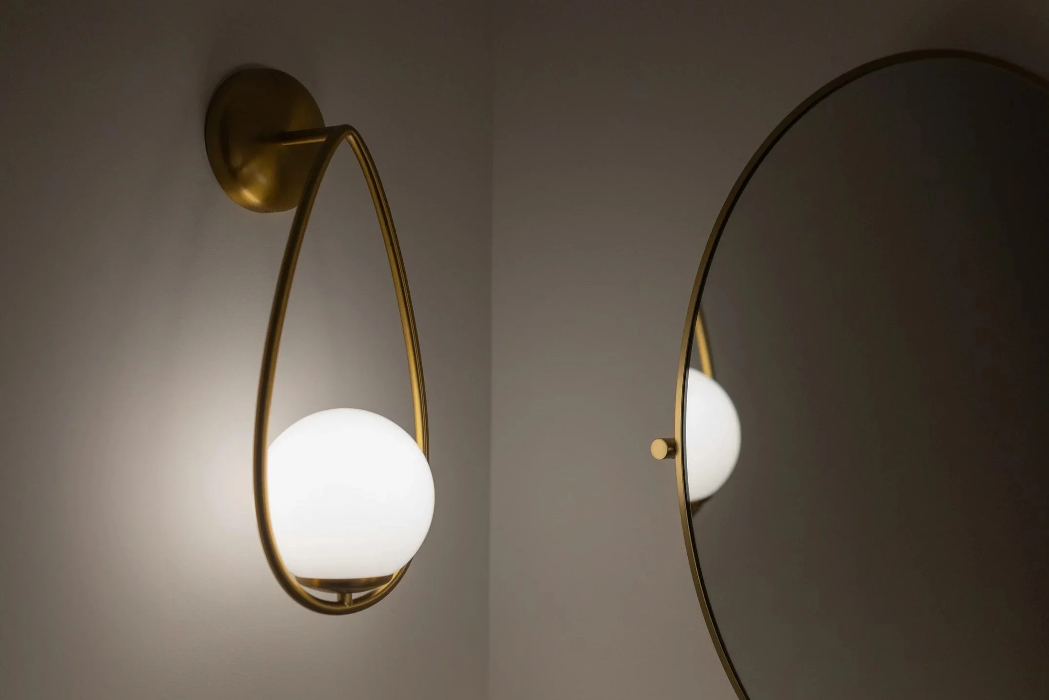 A modern wall-mounted sconce with a round white glass shade and a brass arm, reflected in a round mirror on a white wall.