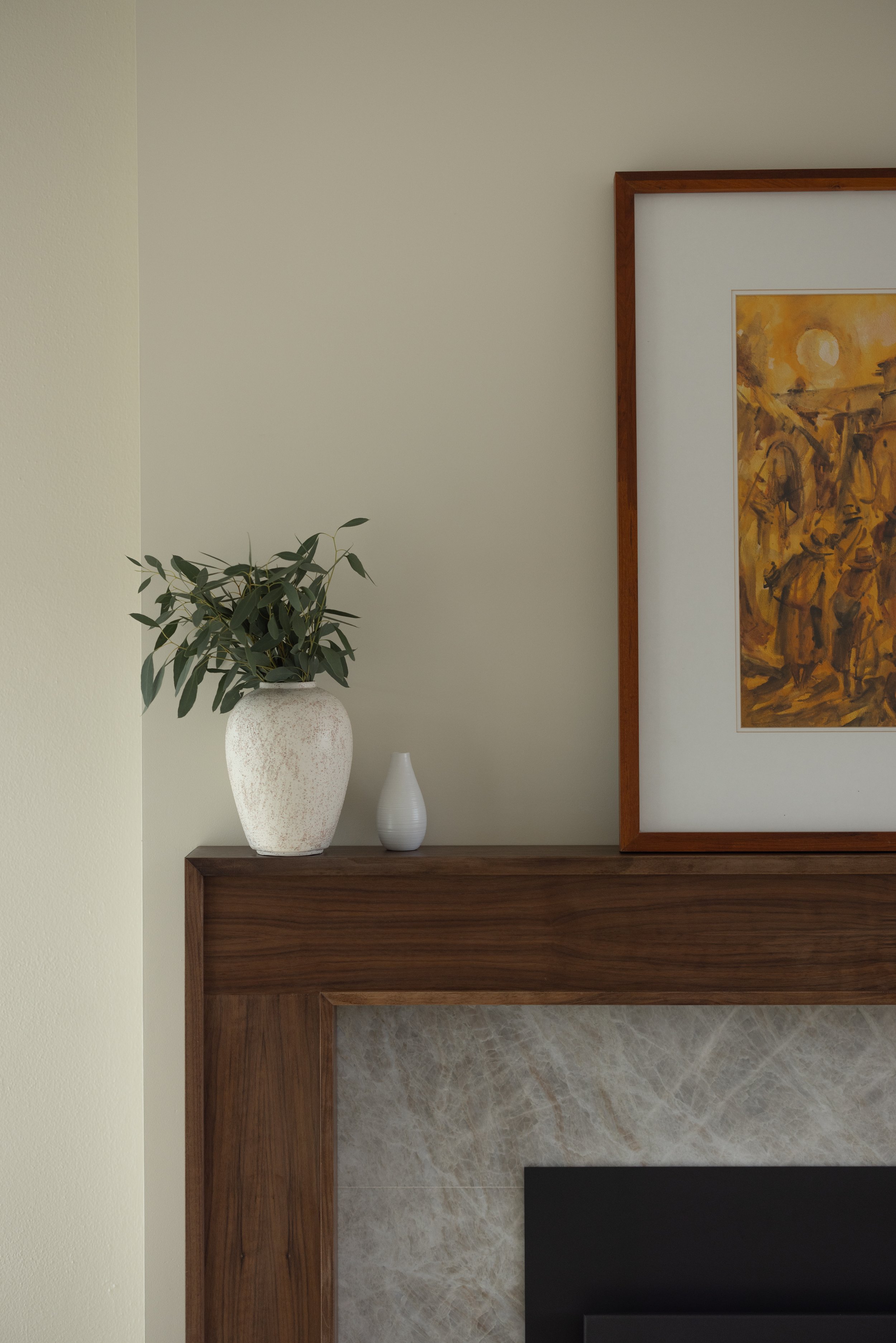 A decorated interior fireplace mantle with a potted green plant and two vases, beside a framed artwork.