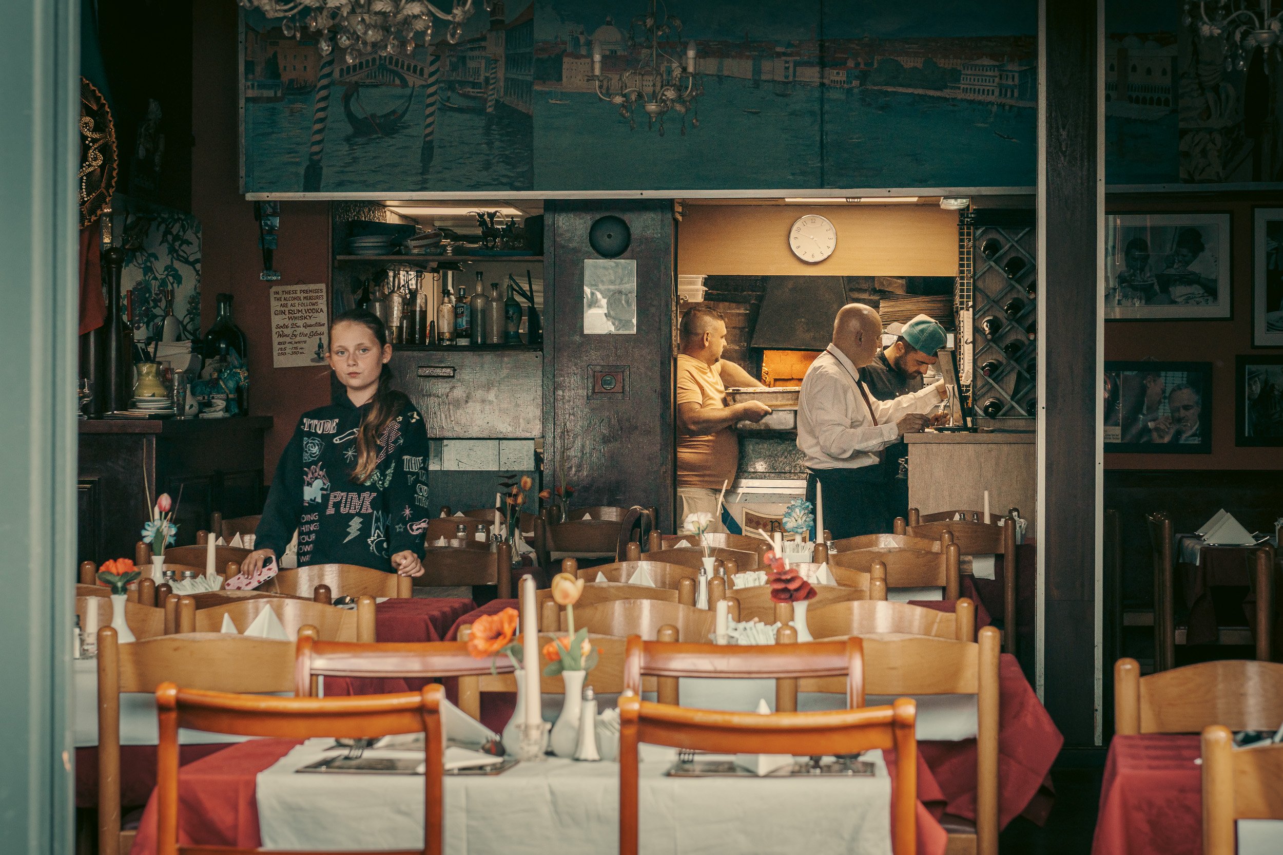 A restaurant interior with tables set with red tablecloths and napkins, and small flower arrangements. There are four people behind the counter, including two chefs and a woman, preparing food. A girl is in the foreground, standing near the tables.