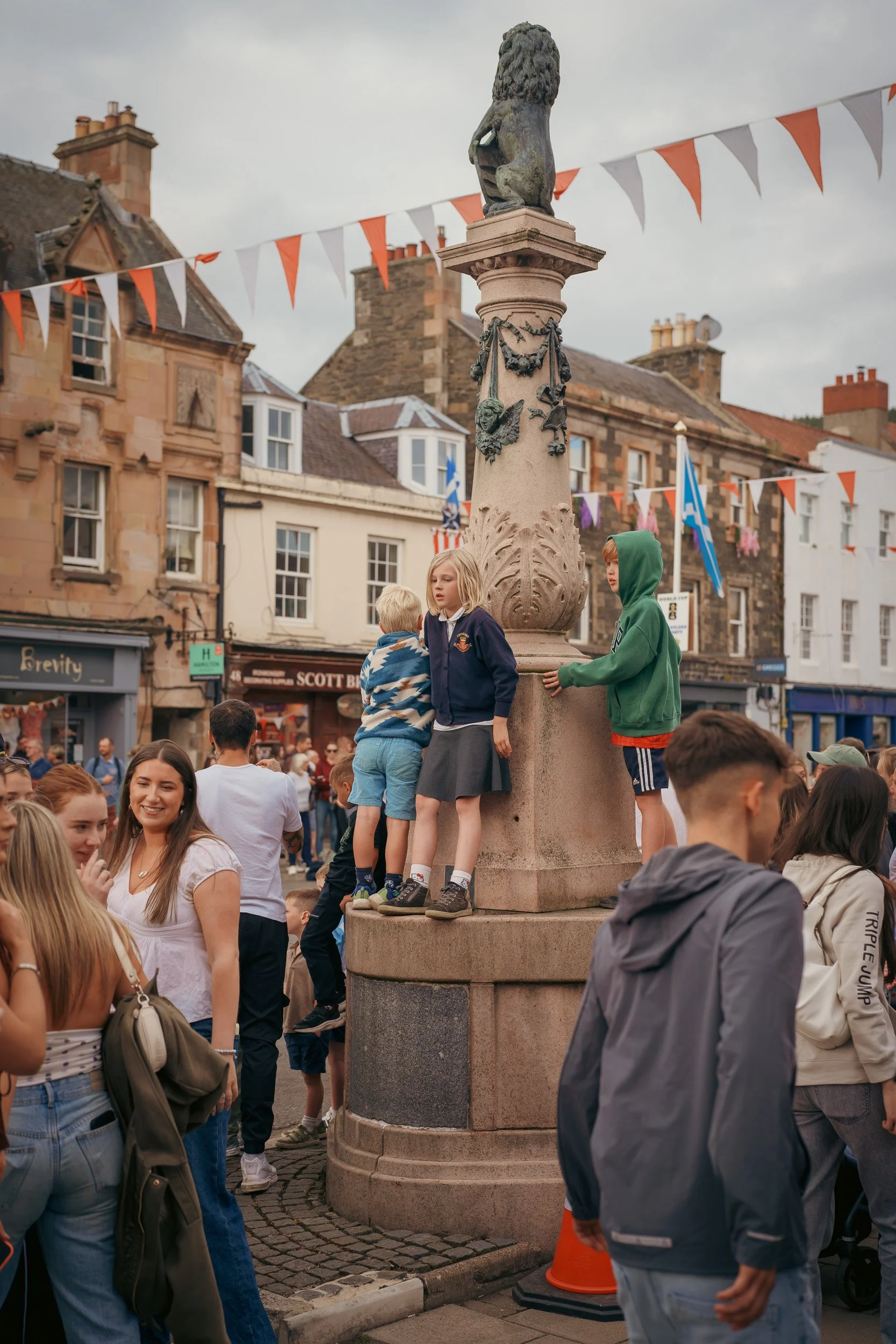 A crowd of people gathered around a stone monument in a town square, with historic buildings and orange and white bunting flags in the background.