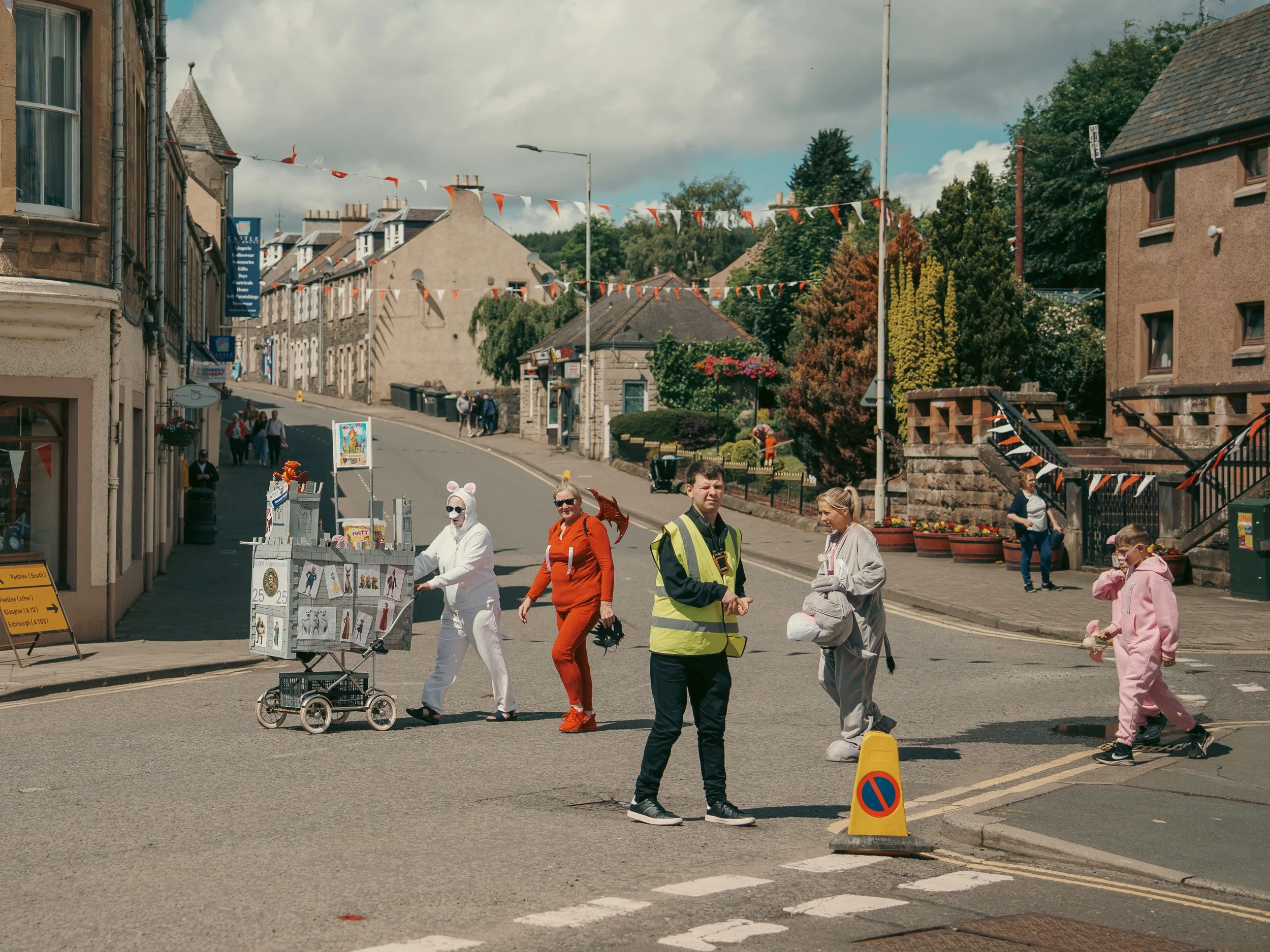People walking on a street decorated with orange and white flags, some in costumes, in what appears to be a festive or parade setting in a small town.