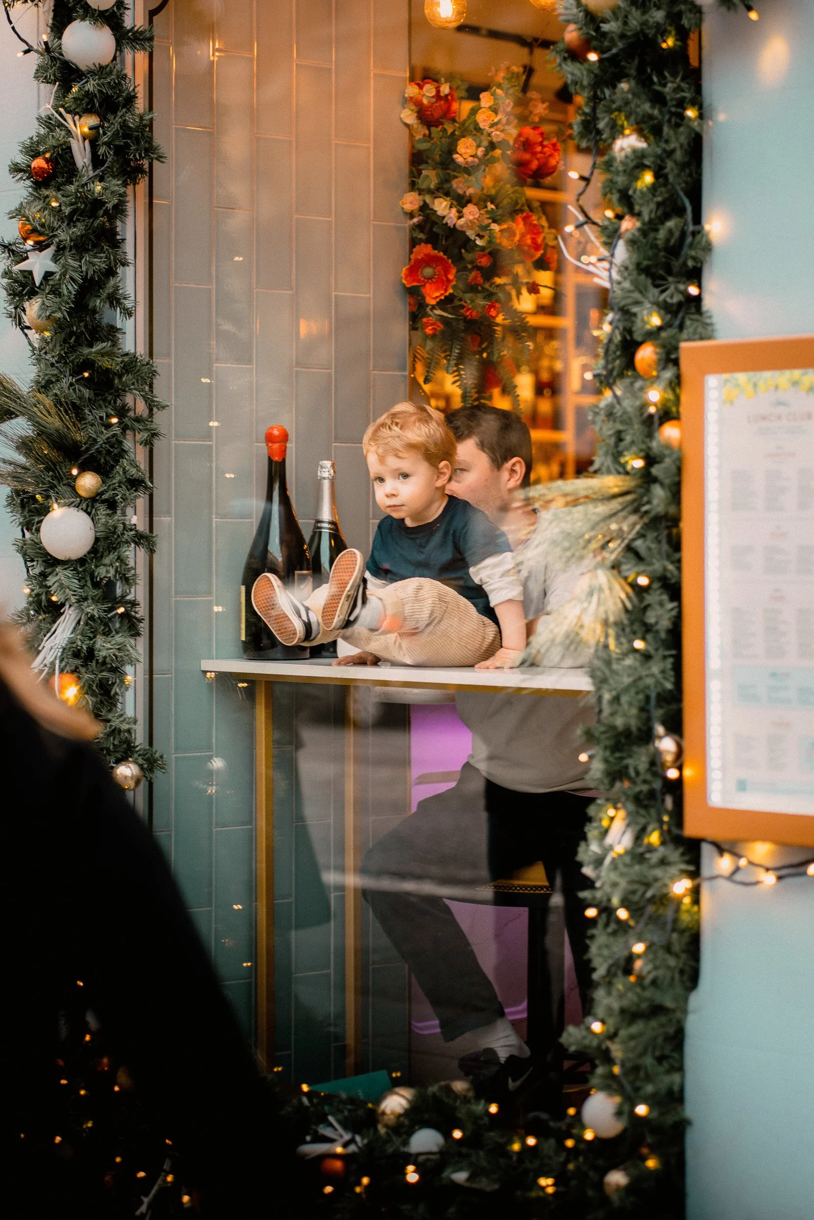 A young boy sitting on a bar counter with a man behind him, framed by decorated Christmas garland and lights in a festive setting.