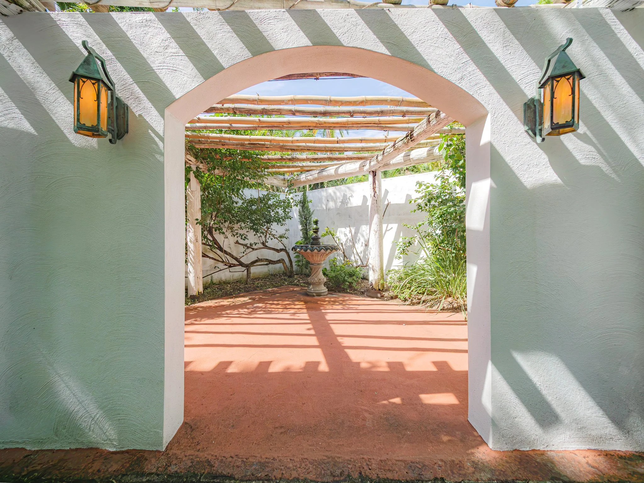 View through an archway into a garden with a fountain, plants, and a bamboo pergola, shaded by a white stucco wall and an overhanging roof with lanterns.