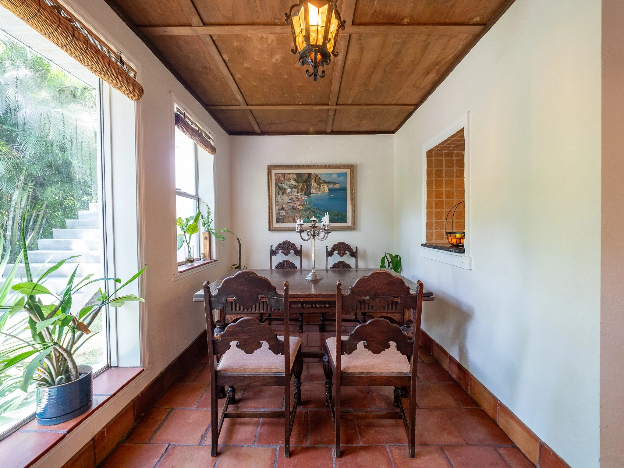 A dining area with a wooden table and four chairs, a chandelier hanging from a wooden ceiling, a framed painting of a coastal landscape on the wall, potted plants on the windowsill, and large windows with blinds providing natural light.