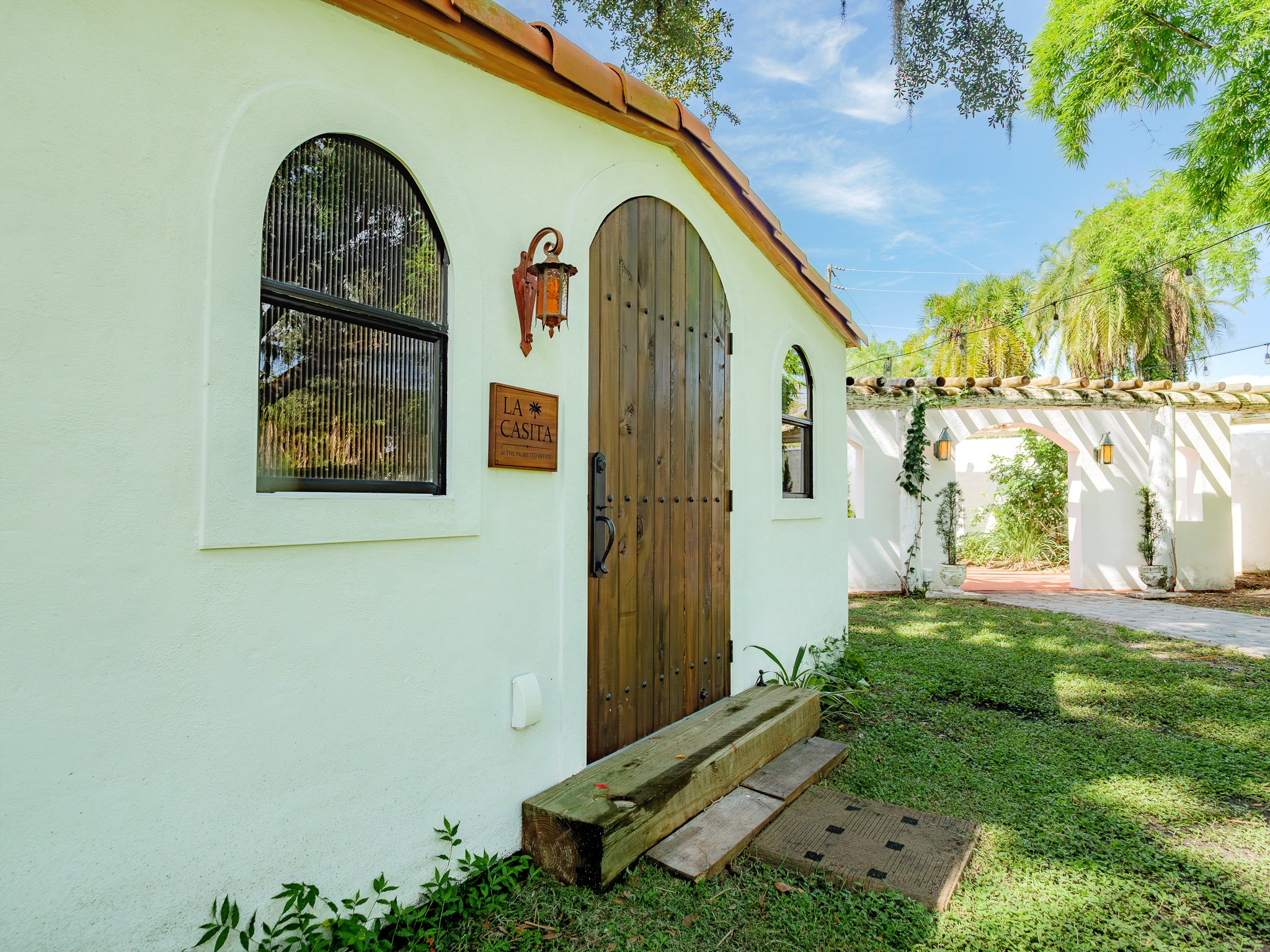 The exterior of a white stucco building with a wooden arched door, a metal-framed window reflecting trees, a lantern-style light fixture, and a sign reading 'La Casita.' Lush green trees, and a clear blue sky.