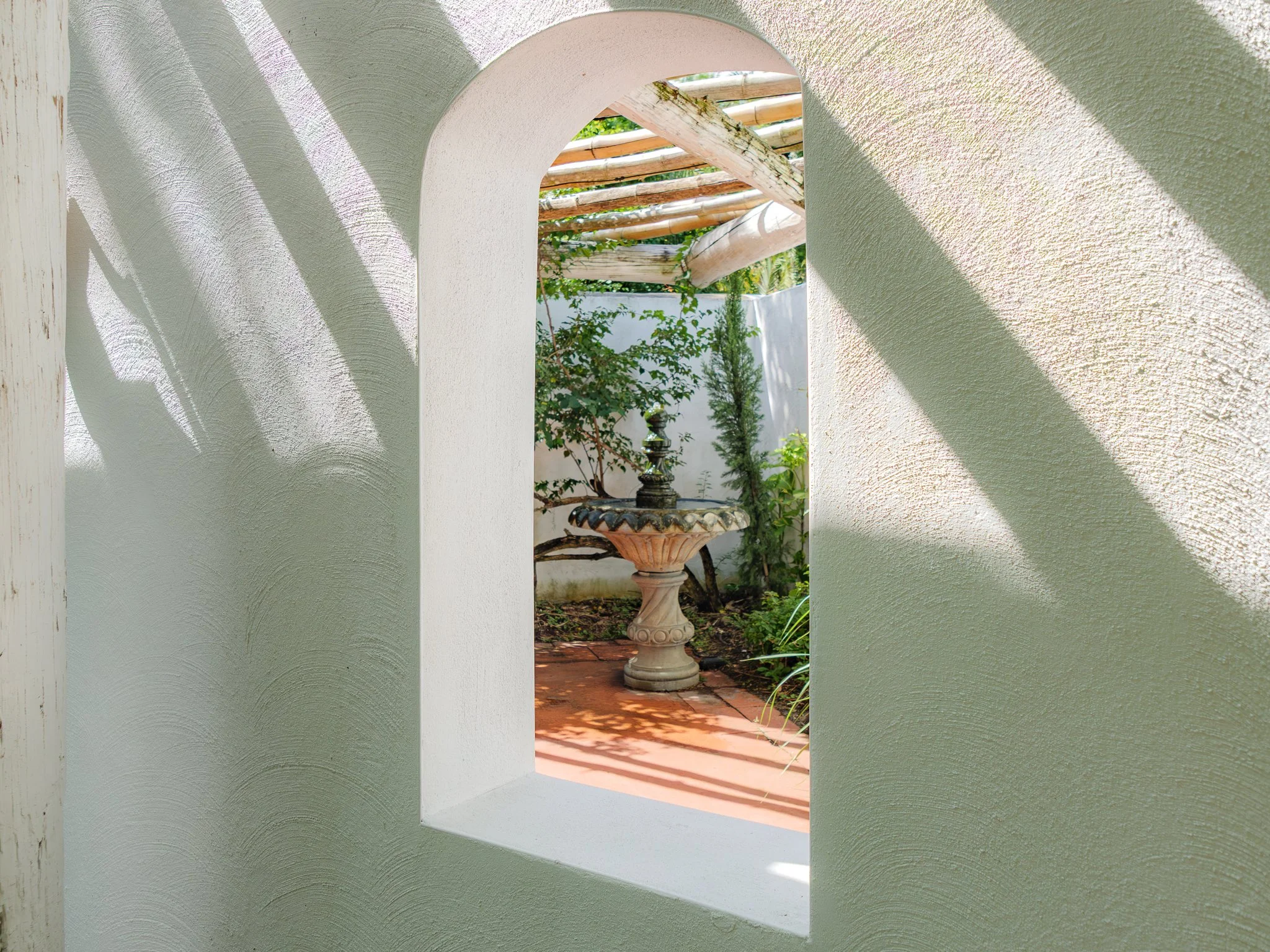 A view through a small arched opening in a wall, showing a decorative fountain in a garden.