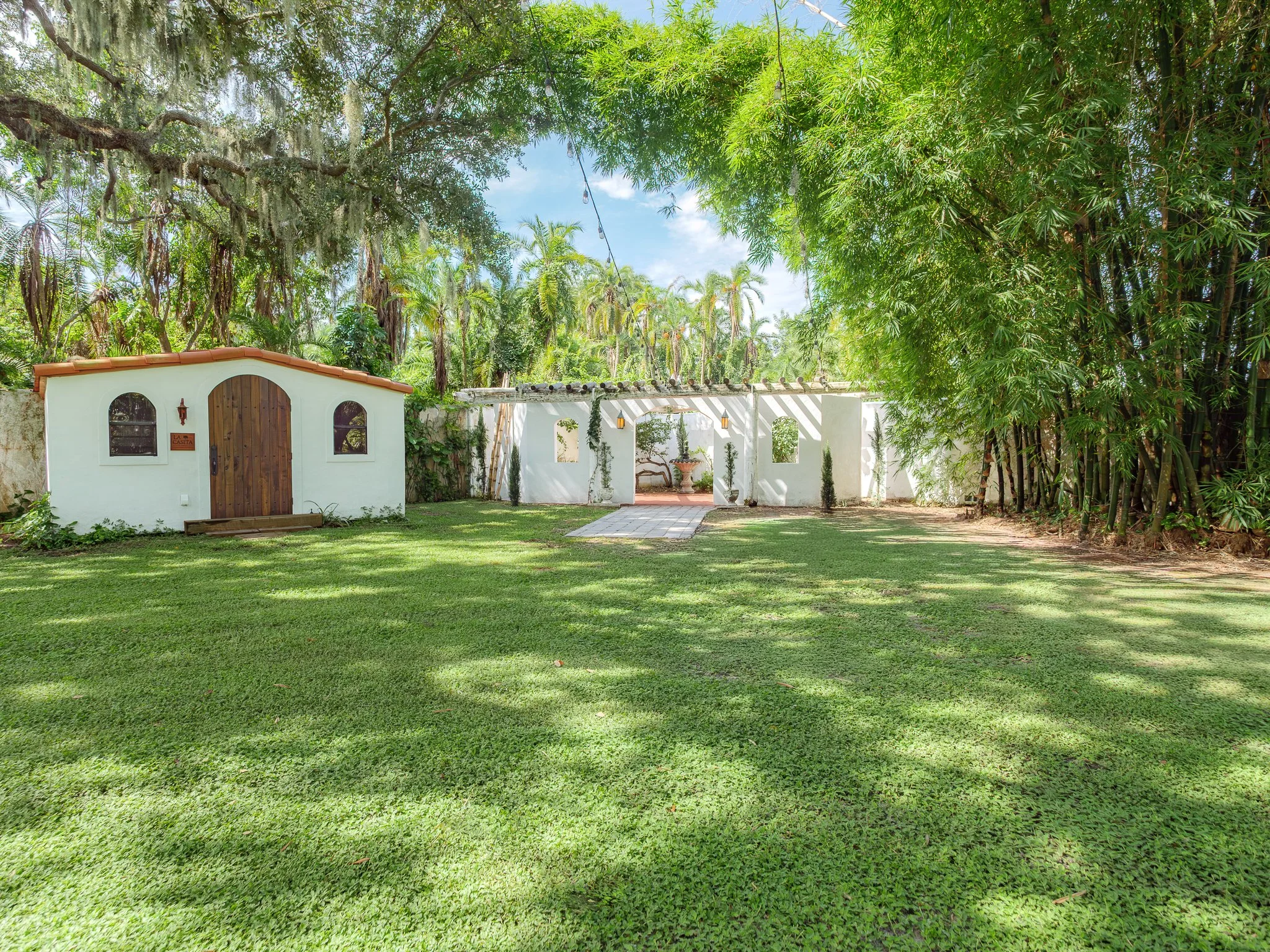 A beautiful backyard garden with lush green grass, large trees, and a white wall with a wooden gate. There are decorative string lights hanging across the yard and small plants along the wall. The sky is blue with some clouds.