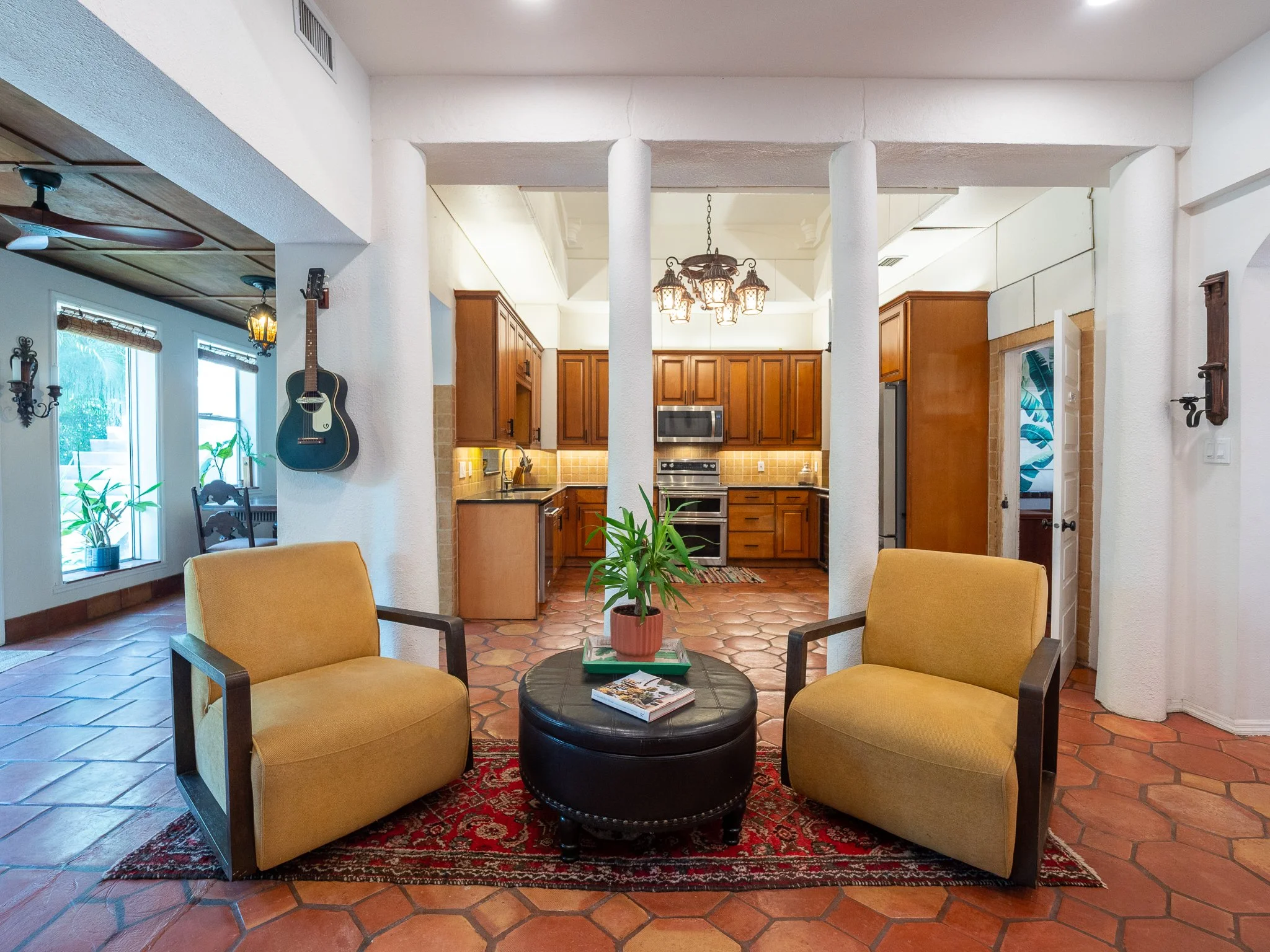 Living room with yellow armchairs, a black round ottoman with magazines on it, potted plant, and a view into the kitchen with wooden cabinets and a hanging chandelier.