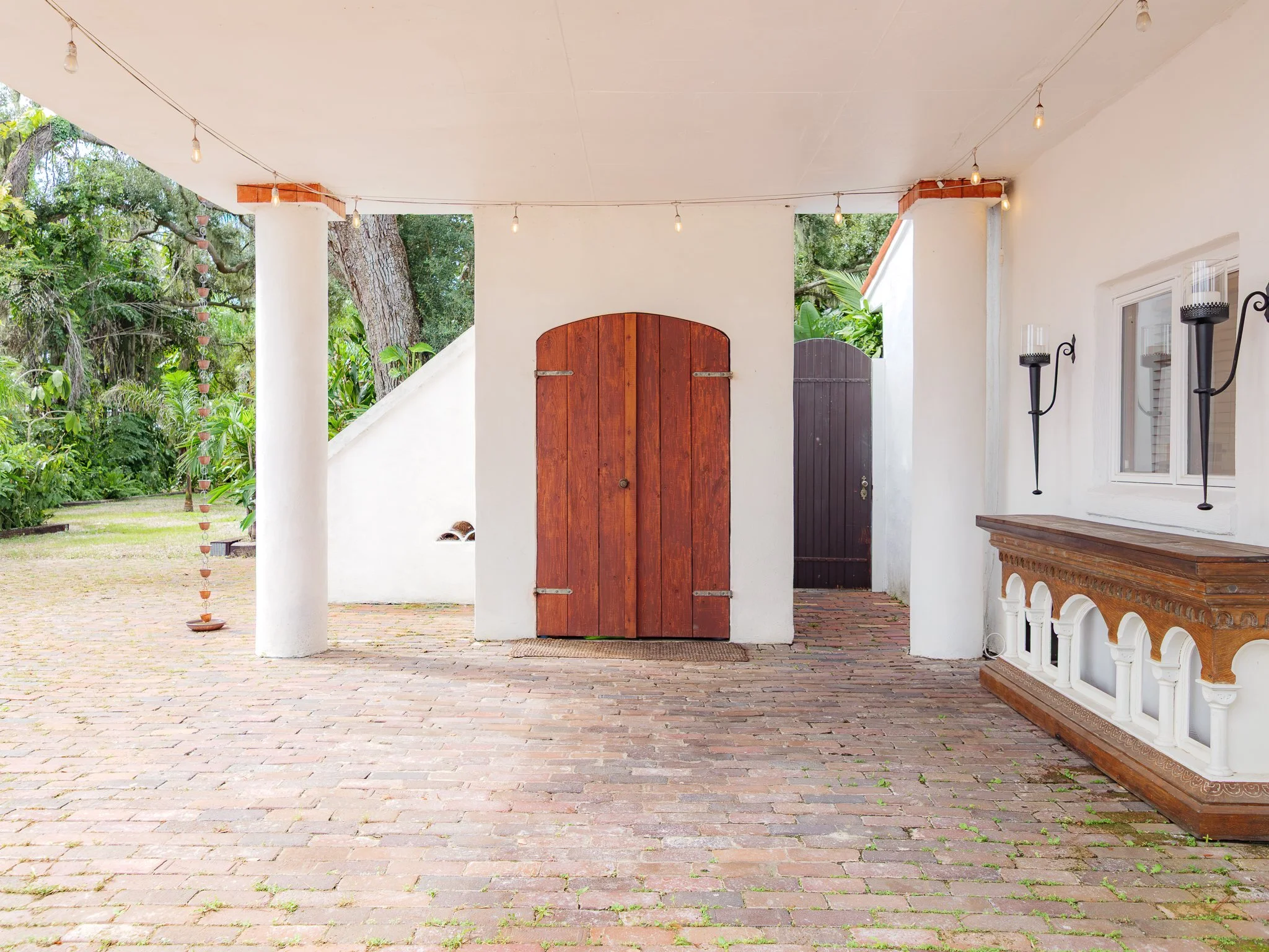 Covered area under balcony with brick flooring, white walls, wooden doors, string lights, and outdoor greenery in the background.