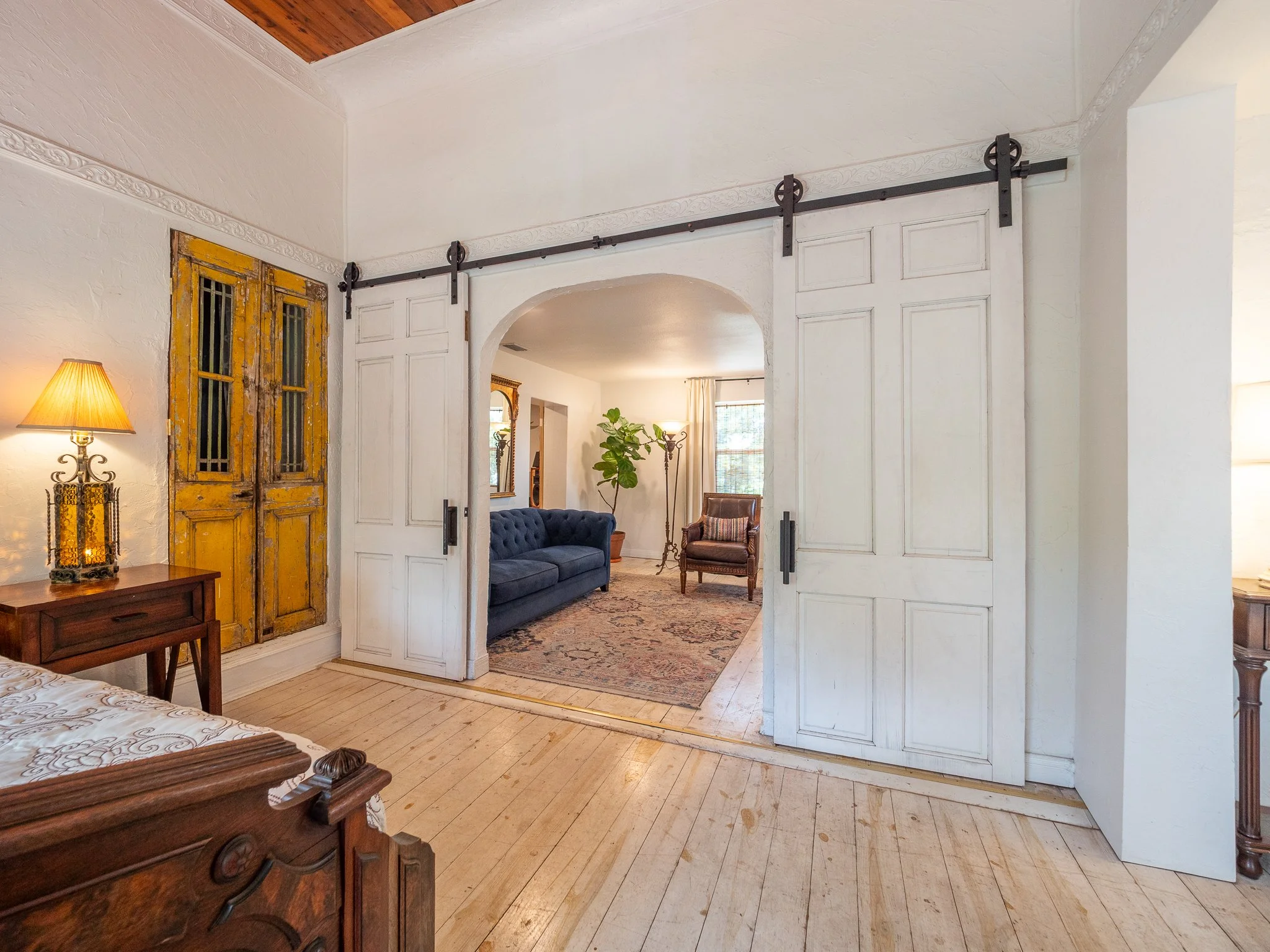 Interior of a house showing a  door with a sliding door mechanism, furniture including a wooden bed with a decorative lamp, a blue sofa, a wooden chest, and a standing lamp. The room has white walls with textured wall moldings, a wooden floor, and se