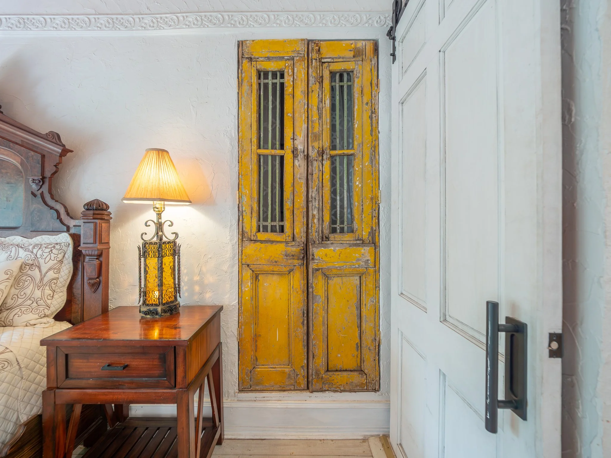A rustic yellow wooden double door with barred windows, set against a white textured wall, with a bedside table and lamp on the left side of the image.