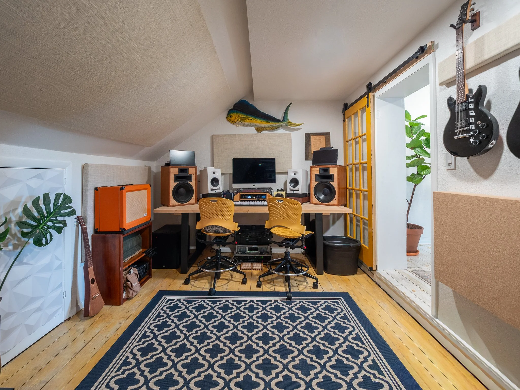 A music studio or office space with a sloped ceiling, featuring a large fish-shaped plush or sculpture on the wall, yellow sliding barn door, plants, orange amplifier, two white speakers, a workspace with a computer and mixing equipment, guitars, and