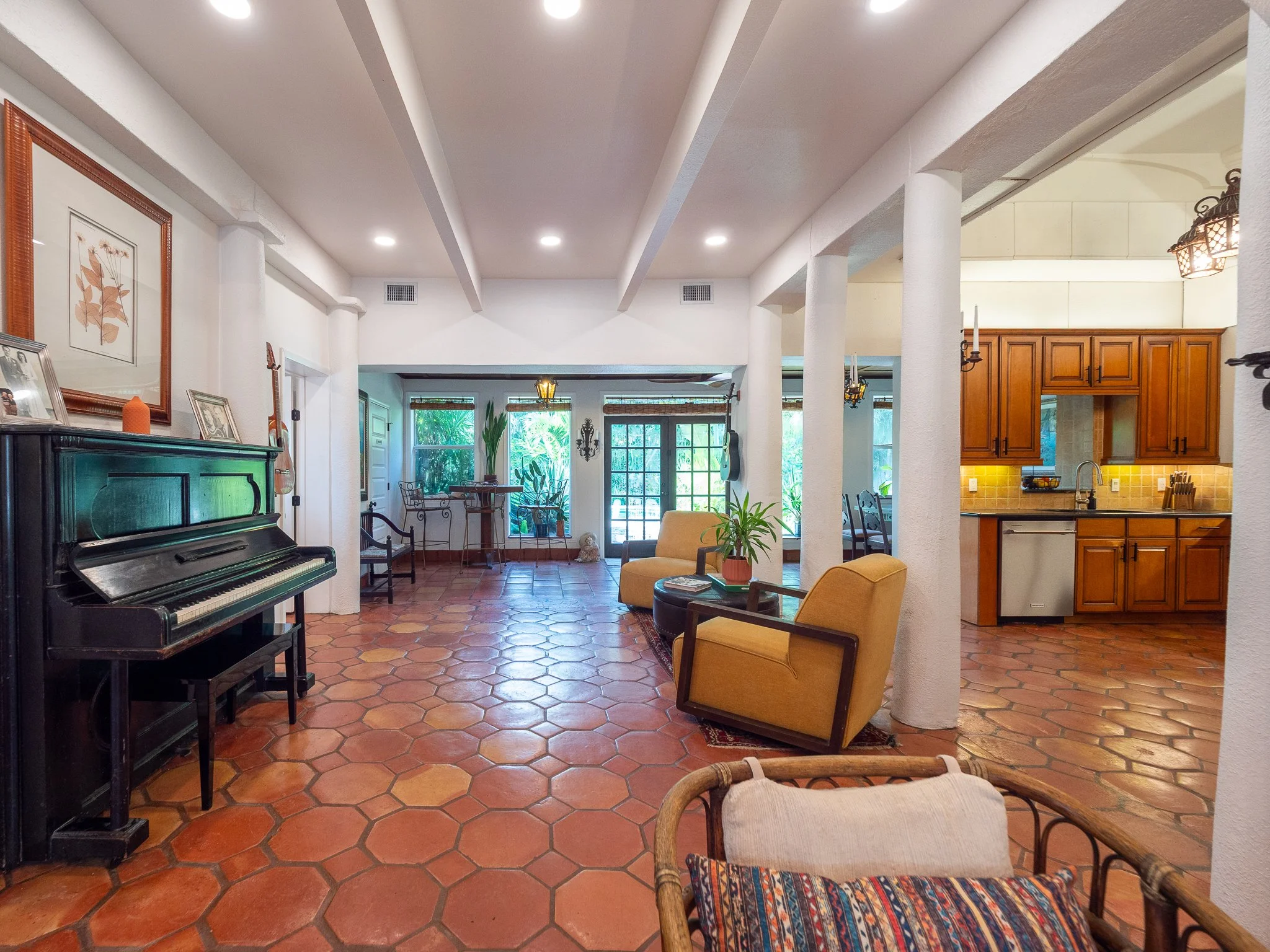 Interior view of a living room with a piano on the left, terracotta tile flooring, a yellow armchair, a wicker chair, and a kitchen area with wooden cabinets on the right, large windows at the back, and plants near the entrance.