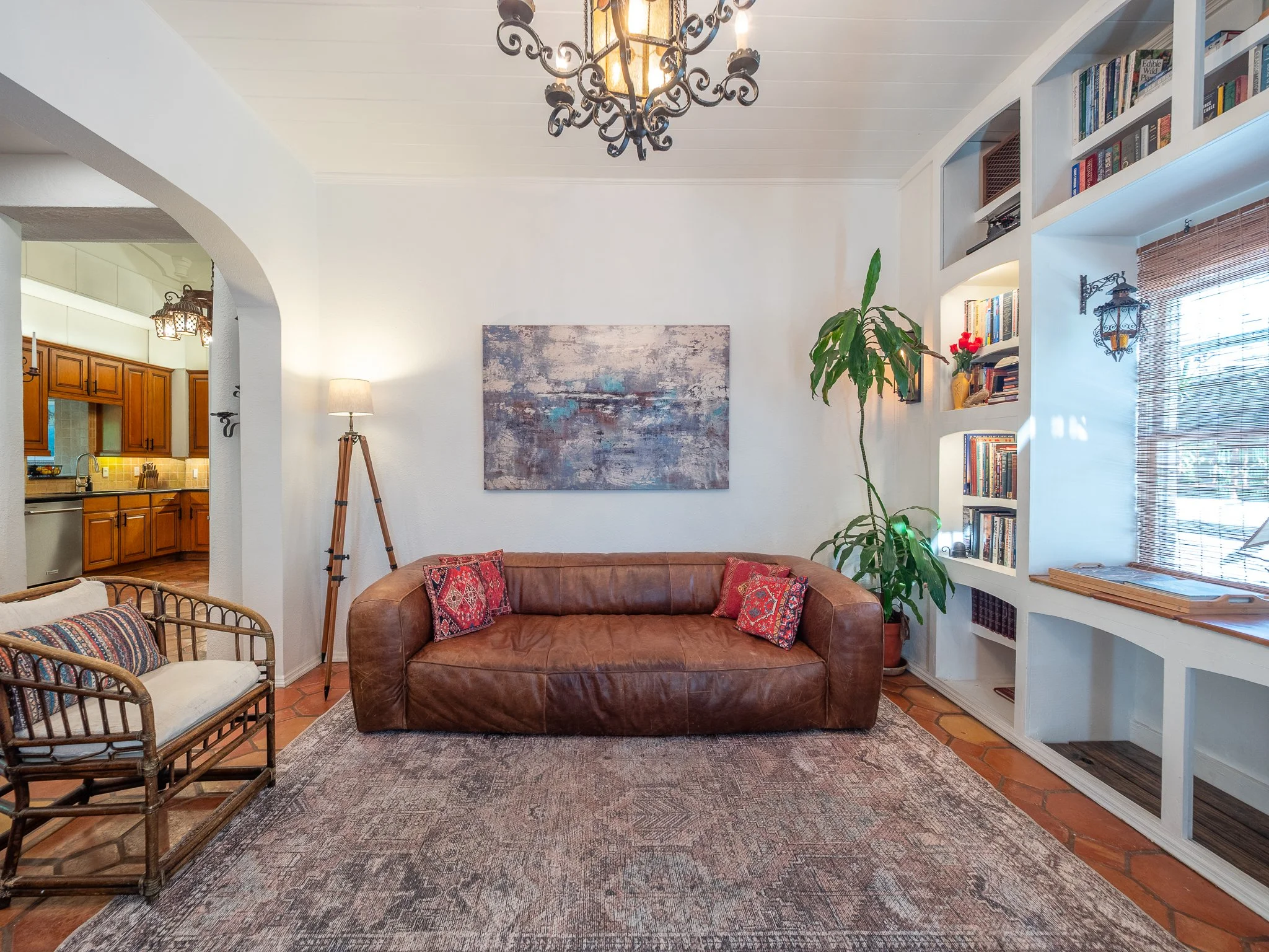 Living room with a leather sofa, patterned cushions, a large abstract painting on the wall, bookshelf built into the wall, potted plant, window with bamboo blinds, chandelier, nearby lamps, and part of the kitchen visible through an arched doorway.
