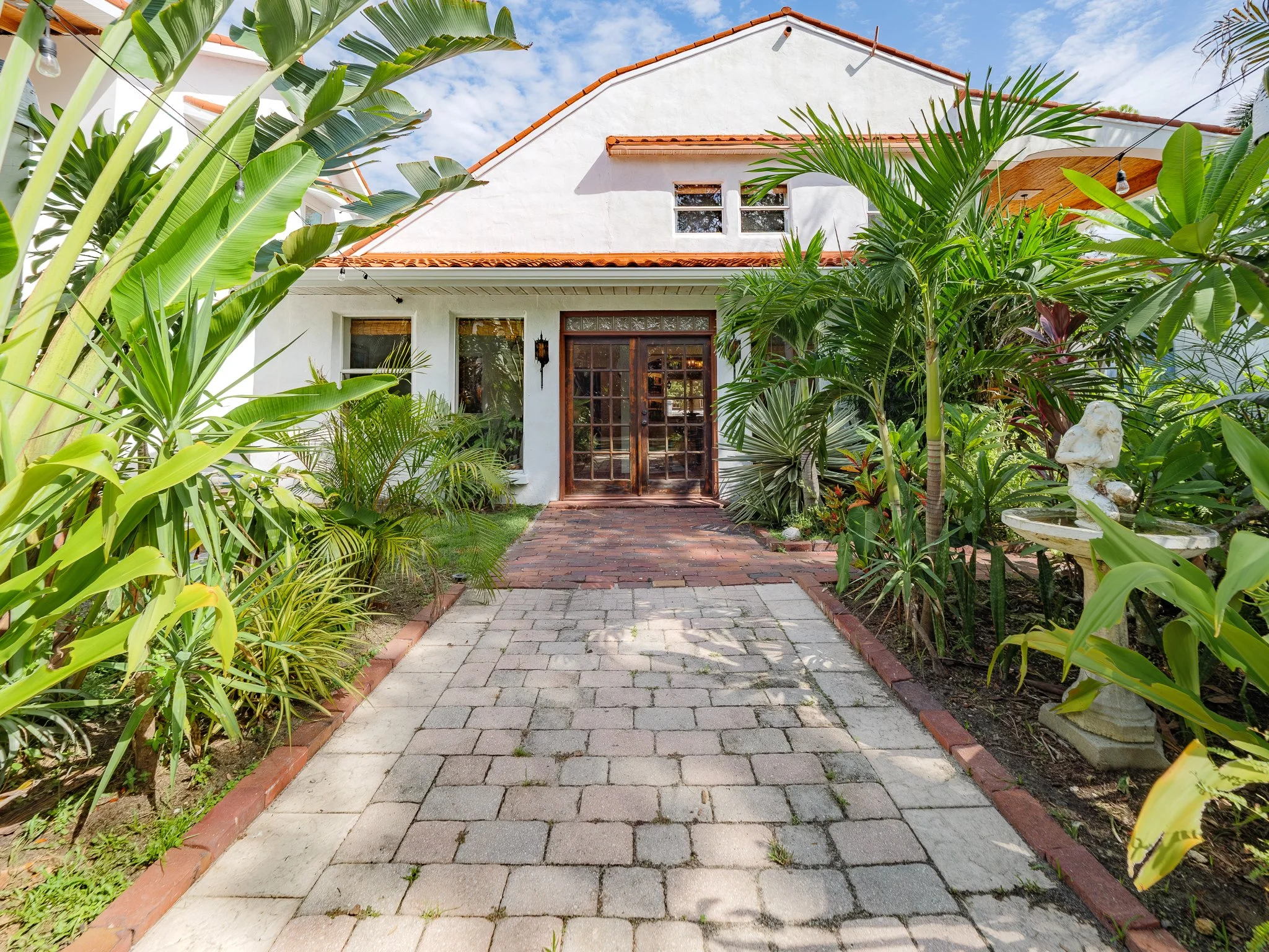 Pathway leading to a Spanish Revival home with a Spanish tile roof, surrounded by lush green tropical plants and trees, under a partly cloudy sky.