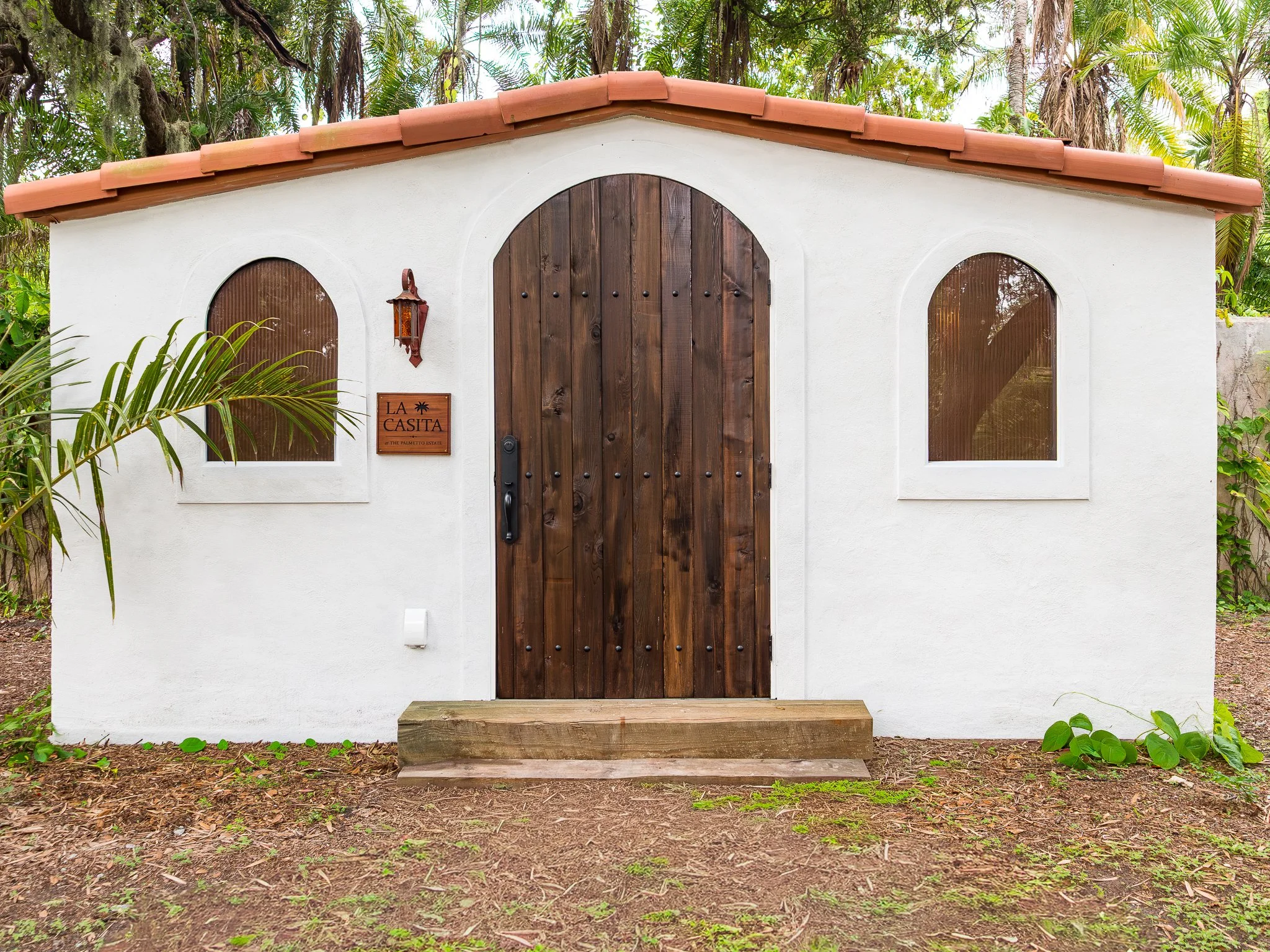 Small white stucco building with a brown wooden arched door and two matching arched windows, surrounded by lush tropical plants and trees.