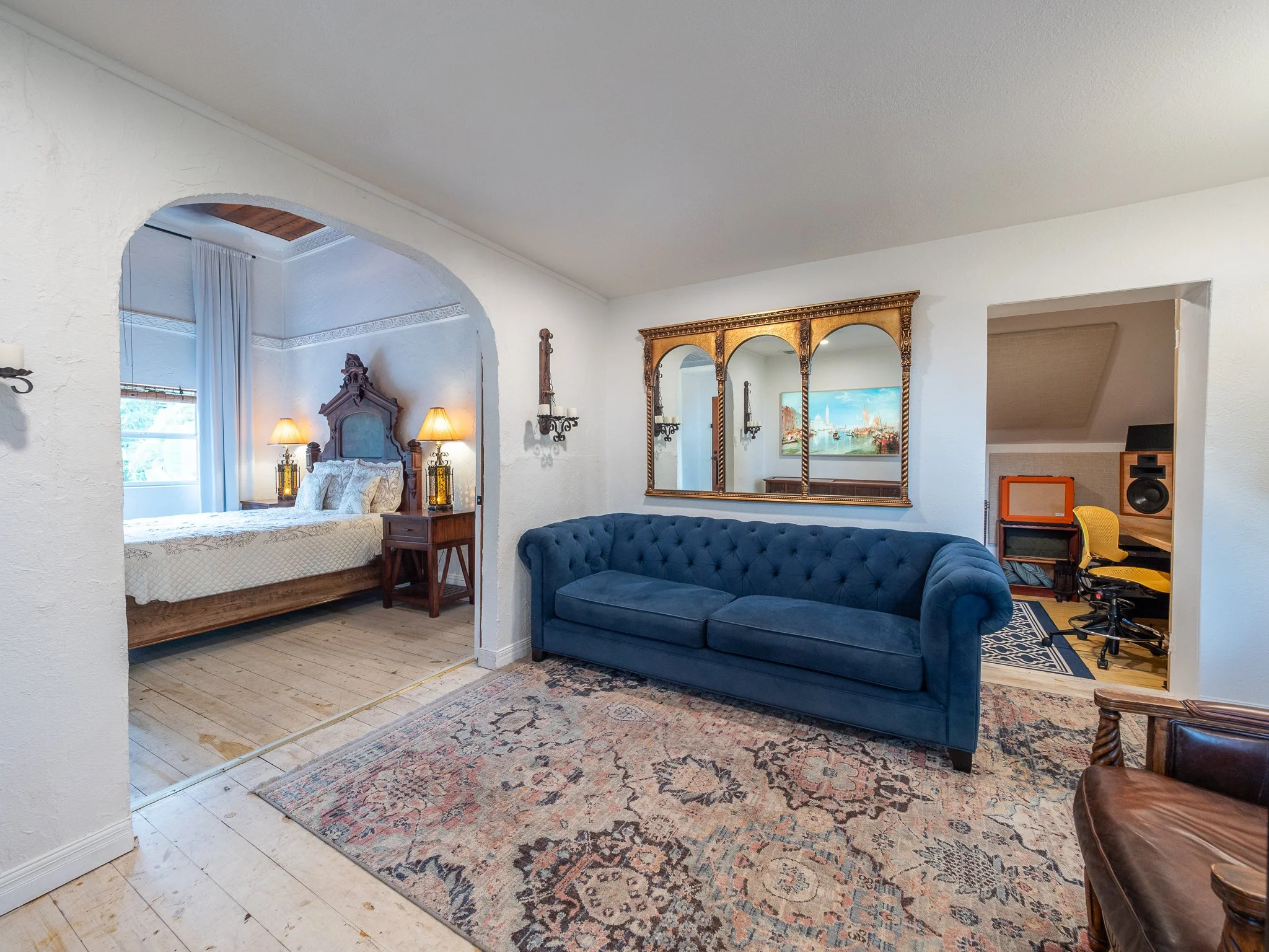 Interior view of a living room and bedroom in a house with white textured walls and wooden floors. The bedroom has a wooden bed with white bedding and bedside lamps. The living room features a large ornate gold-framed mirror, a blue tufted sofa, a co