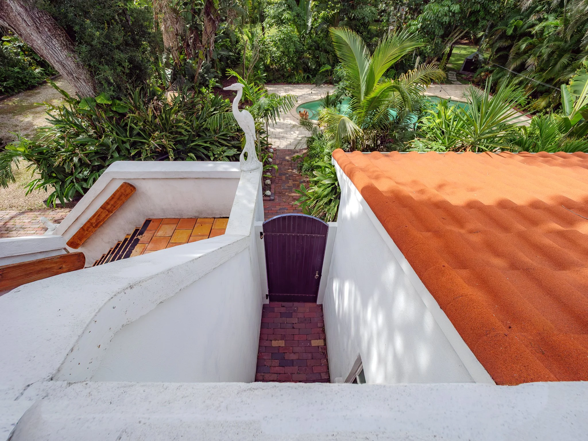 View from a white balcony with roof tiles, overlooking a landscaped backyard with various lush green plants and trees, a brick pathway, a small pool, and a wooden heron sculpture.