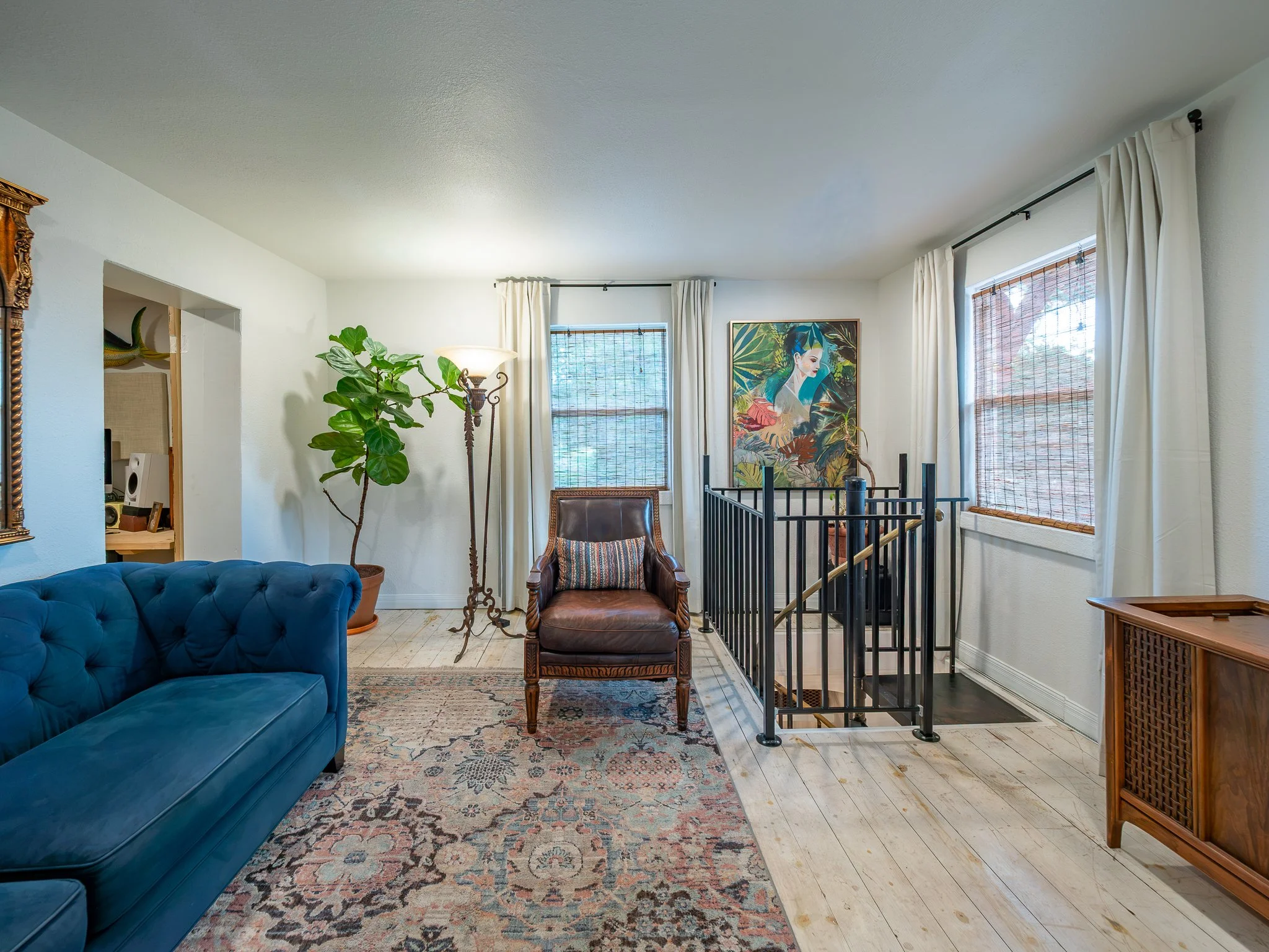 Living room with a blue velvet couch, a vintage armchair, a large potted ficus plant, a patterned area rug, a wooden side table, a tall floor lamp, two windows with white curtains, a colorful art print, and a staircase.
