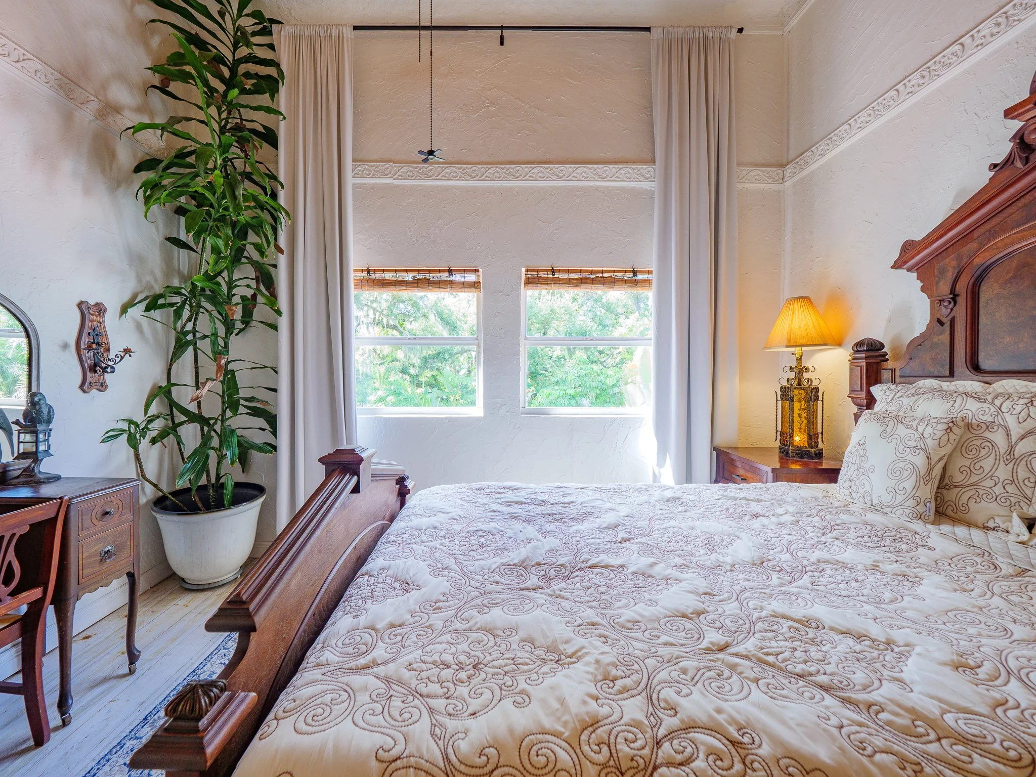 Interior of a bedroom with a wooden bed, an ornate wooden headboard, a nightstand with a lamp, a large potted plant, and a large window with blinds