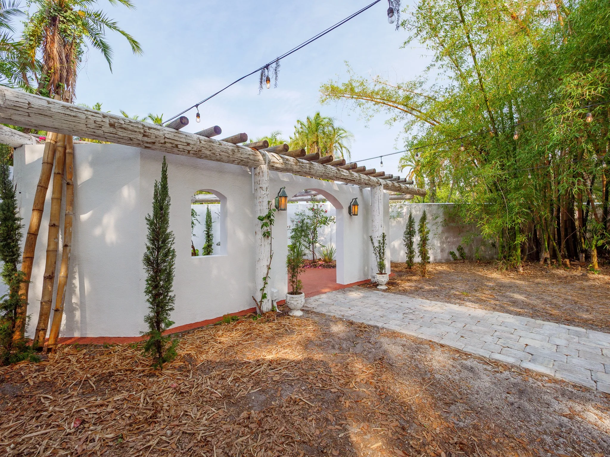 A backyard with a white stucco wall, potted plants, and tall trees. String lights hang above, and the ground is covered with leaves and paving stones.