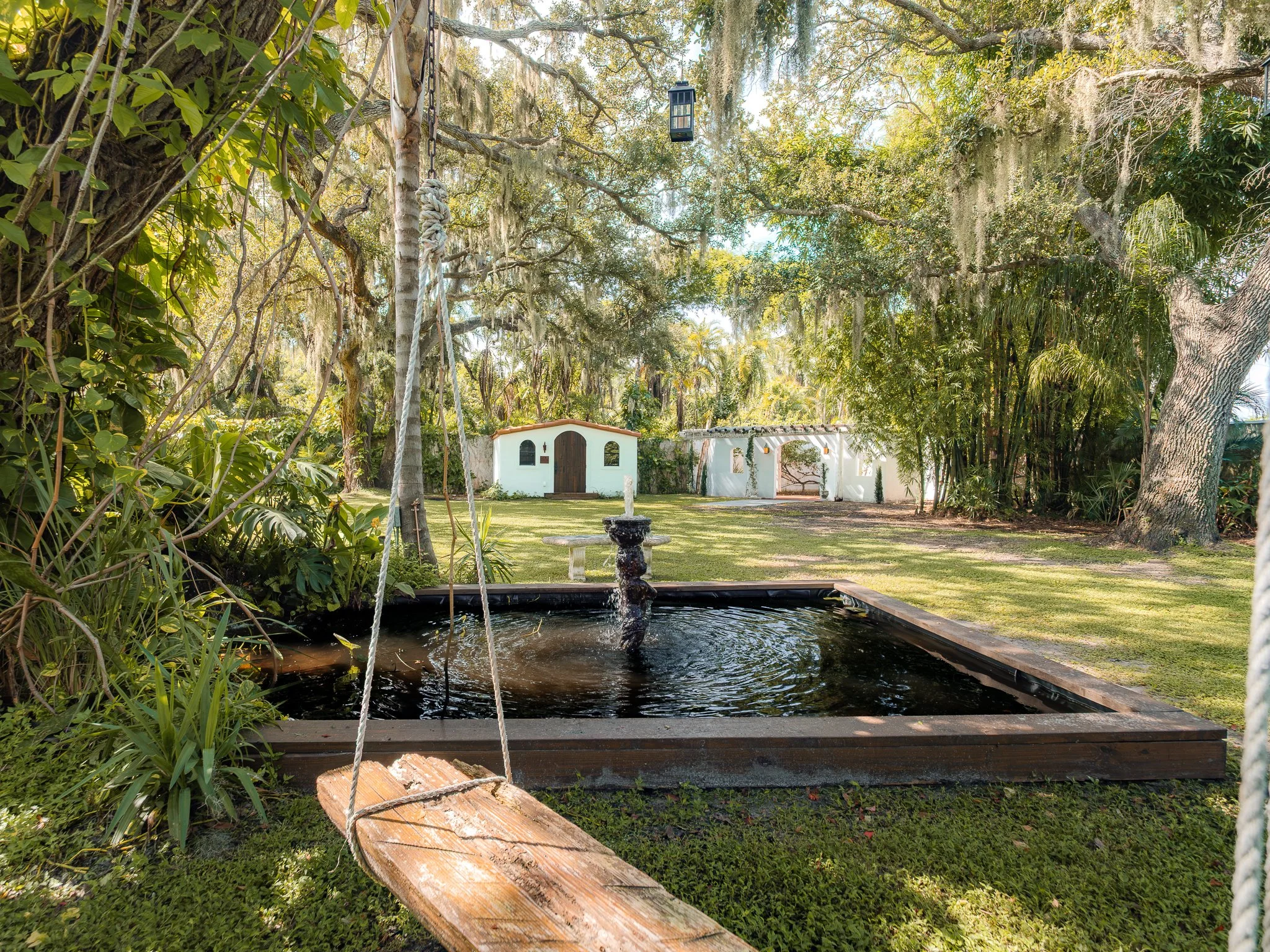 A large backyard with koi pond, a wooden swing, lush trees with Spanish moss, a white Casita, and a Spanish archway in the background.