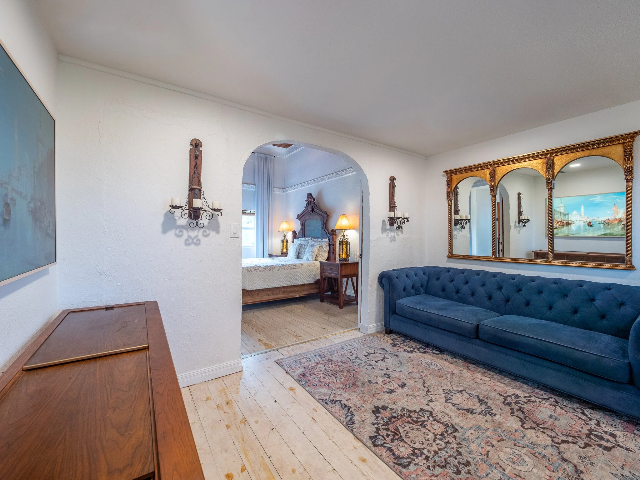 Interior of a living room with a blue velvet sofa, a large ornate gold-framed mirror, wall sconces with candles, and a doorway leading to a bedroom with a ceiling fan, lamps, and a bed.