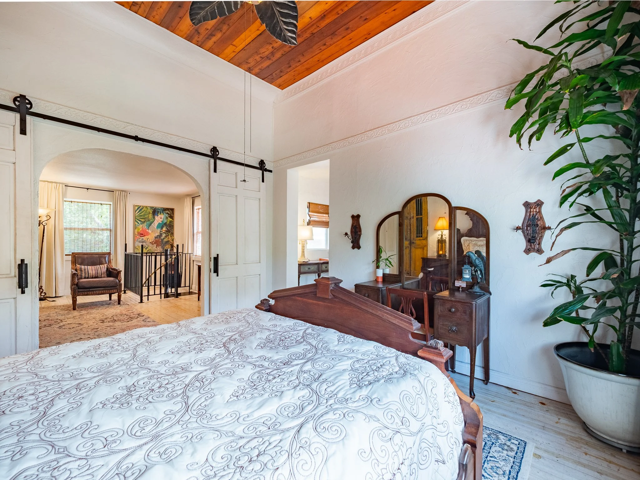 A cozy bedroom with a wooden ceiling and a white textured wall. There is a large bed with white bedding with intricate embroidery, a wooden headboard, and matching bedside tables. A large green potted plant is near the bed. Behind the wall, a sitting