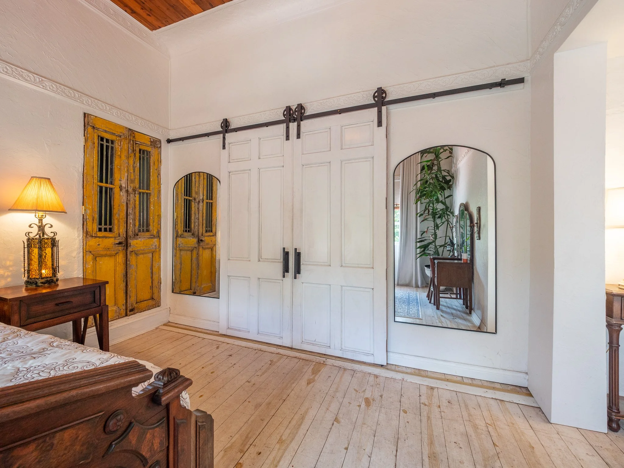 A bedroom with a wooden bed frame, a bedside table with a lamp, a decorative mirror, and a wardrobe with white paneled doors. There are two mirrors on the wall, a tall plant, and a sliding barn door on a black metal track.