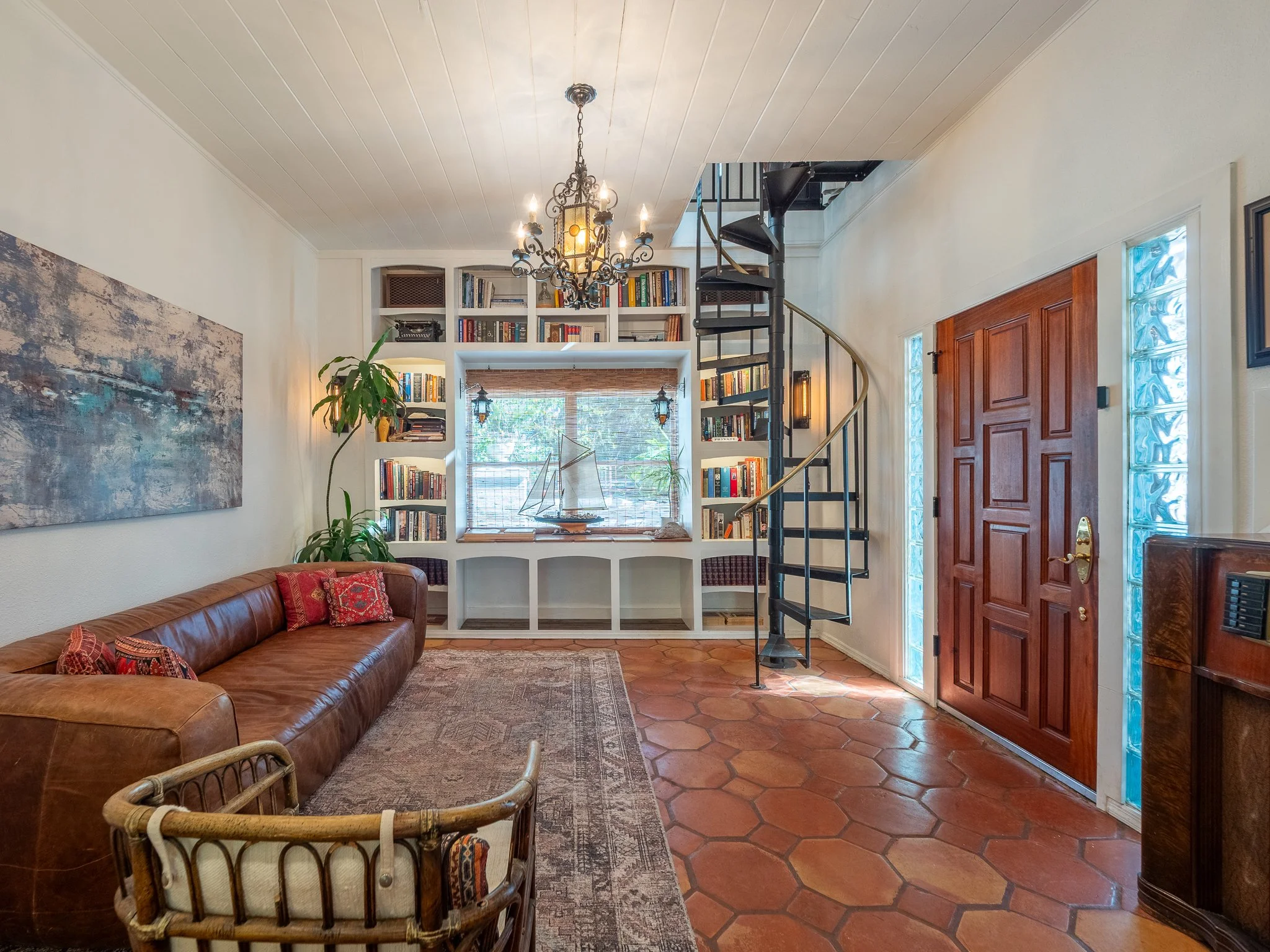 A cozy living room with a brown leather sofa covered with decorative pillows, a patterned area rug, a white built-in bookshelf filled with books, a large window with bamboo blinds, a vintage sailboat model on the windowsill, a chandelier hanging from