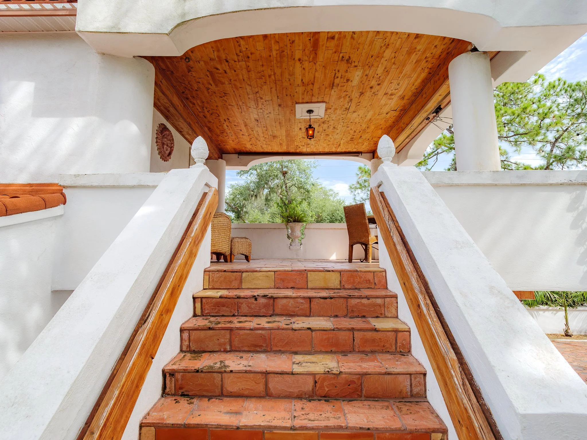 Outdoor staircase leading to a covered patio area with reddish-brown brick steps, wooden ceiling, white walls, and surrounded by green trees and blue sky.