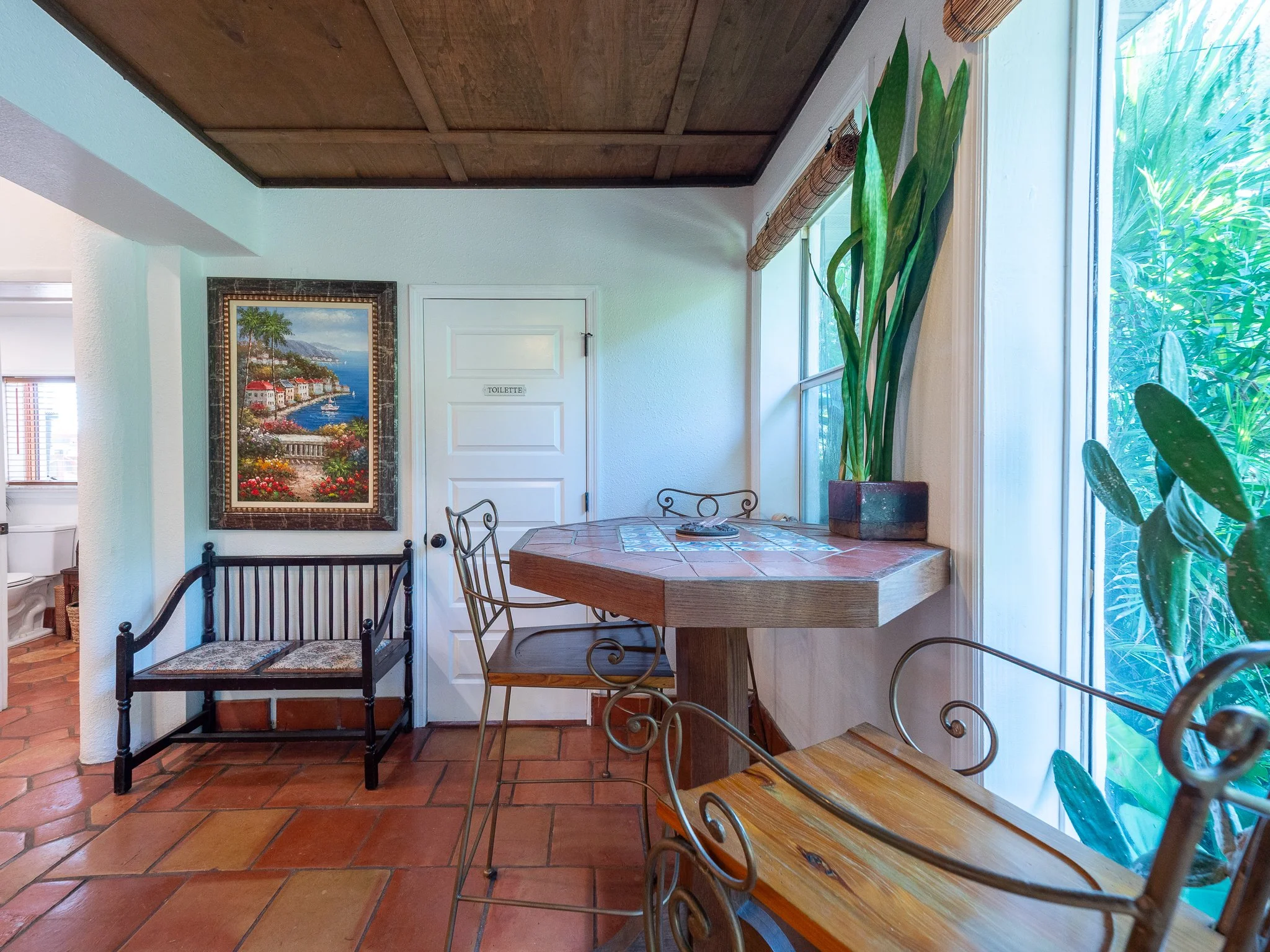 Corner of a room with a white door labeled 'Toilette', a wall with a painting of a harbor scene, a large potted plant, a wooden table, and a window with bamboo blinds.