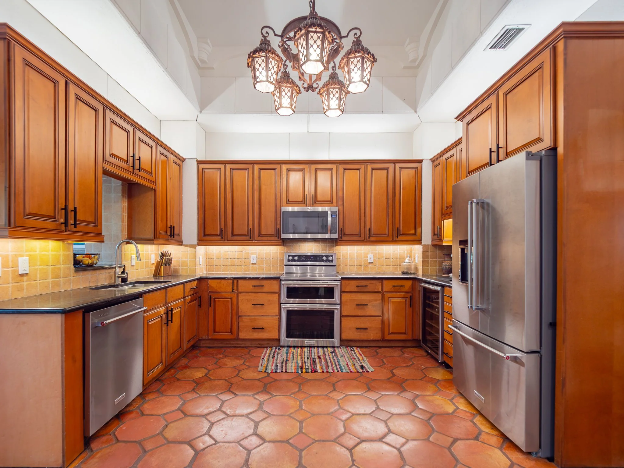 Kitchen with wooden cabinets, stainless steel appliances, a colorful striped rug, and a chandelier with multiple lantern-style lights hanging from the ceiling.