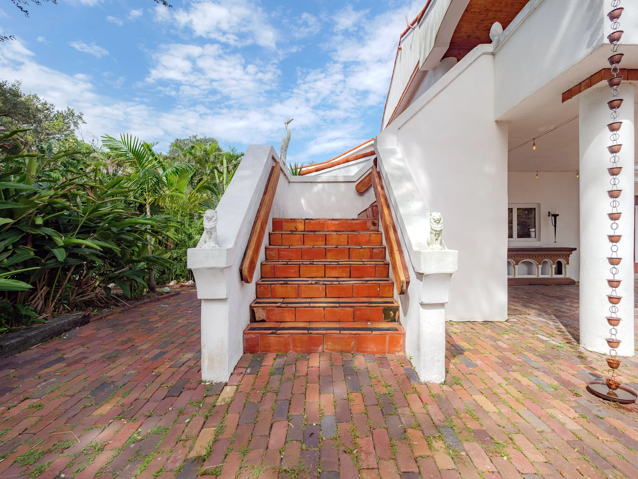 Outdoor scene with brick stairs leading up to a white building with decorative lions at the base of the stairs, surrounded by lush greenery and a partly cloudy sky.