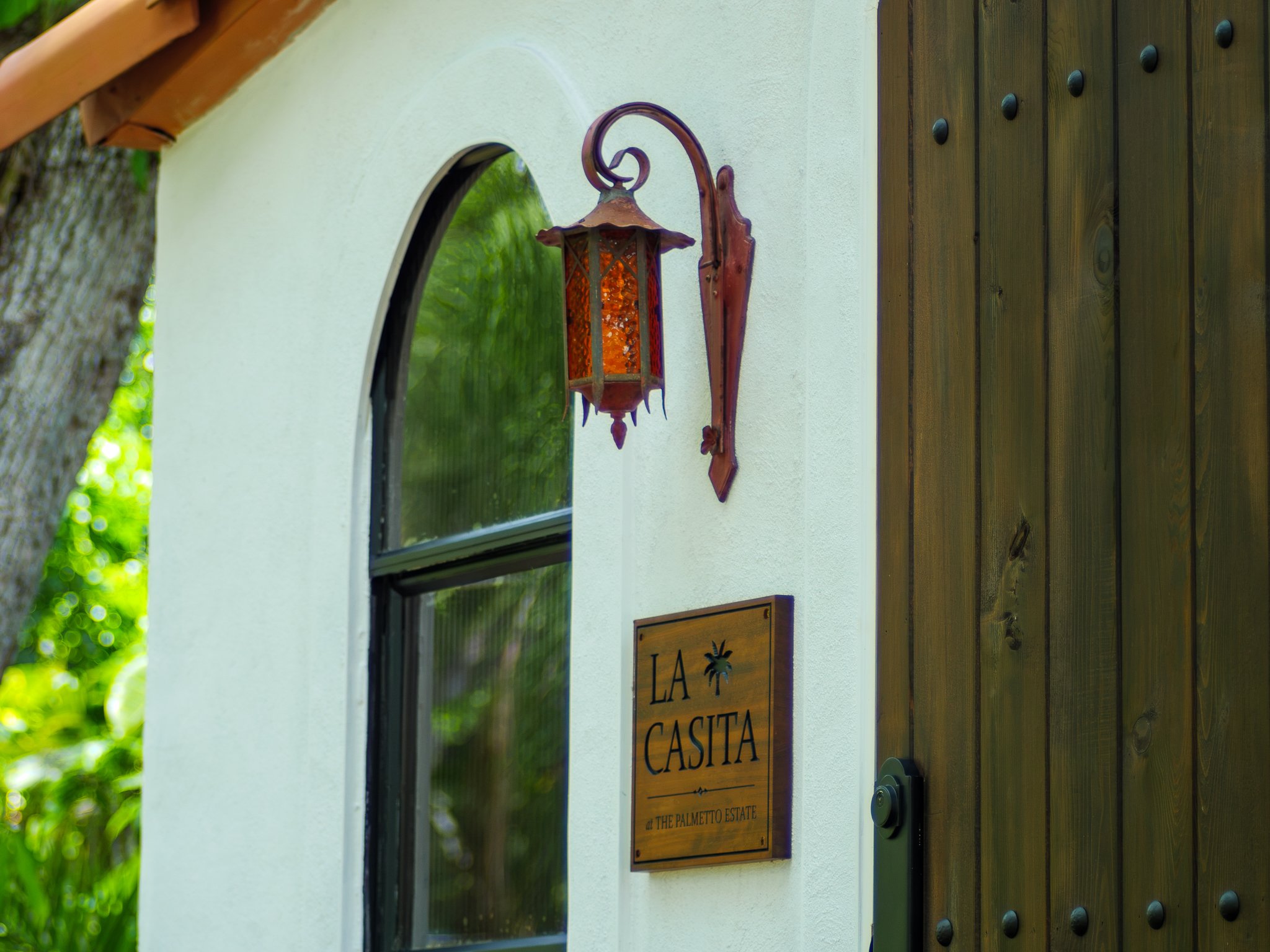 A wooden sign that reads 'La Casita at The Palmetto Estate,' mounted on a white stucco wall with an arched window, outdoor lights, and greenery in the background.