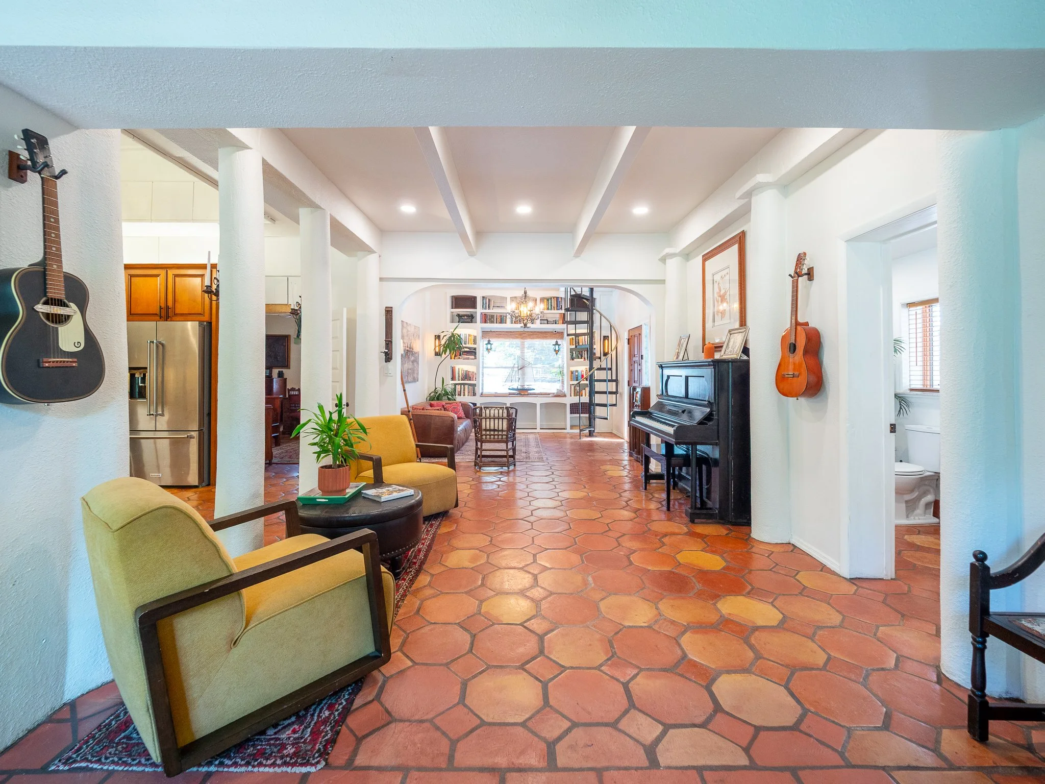Interior of a living room with hexagonal terracotta floor tiles, white textured walls, and a ceiling with exposed beams. There are guitars hanging on the walls, a black upright piano with framed pictures on top, a yellow armchair, a small wooden tabl