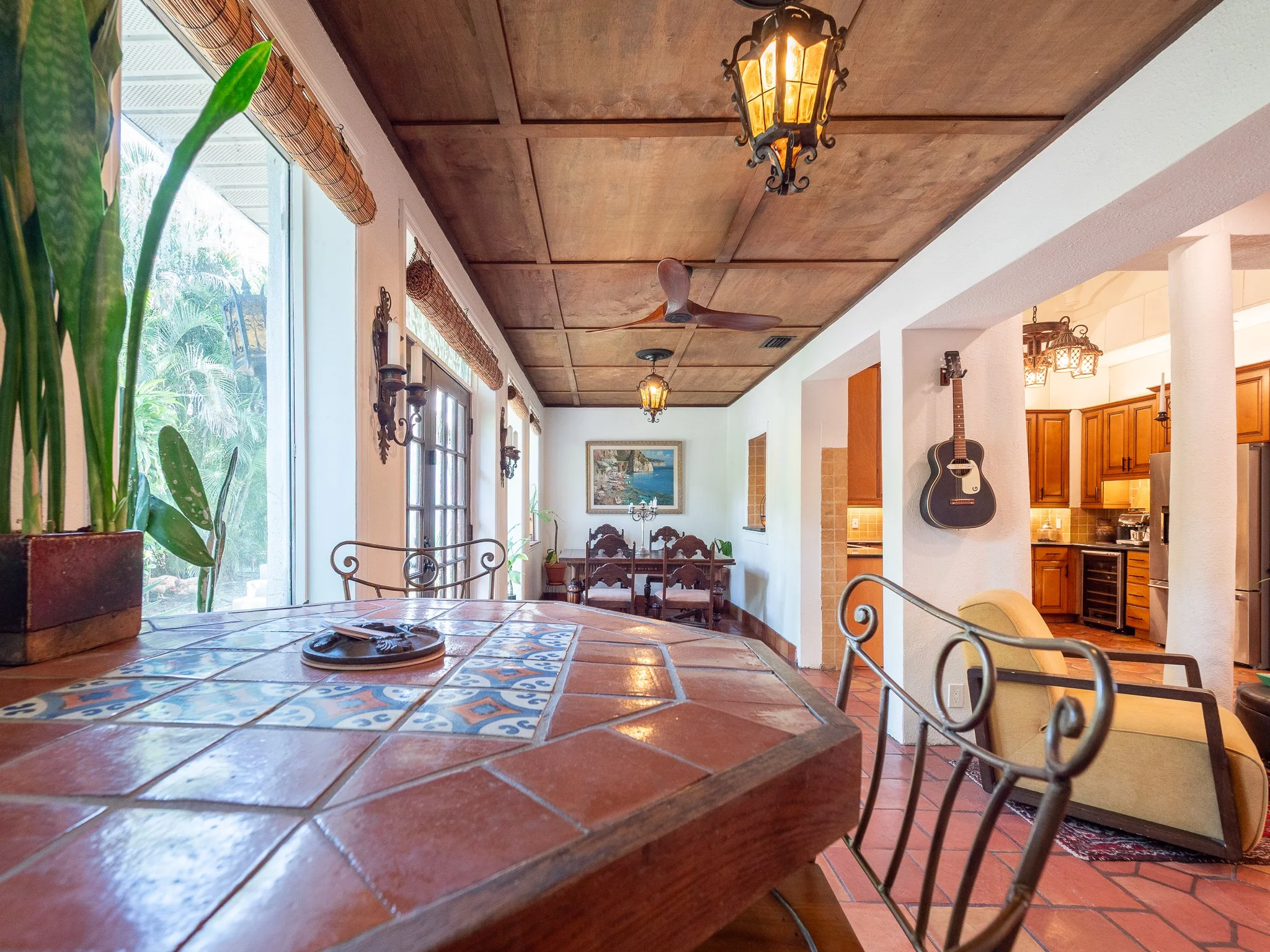 View of a cozy kitchen and dining area with a wooden ceiling, hanging lantern-style lights, a ceiling fan, a guitar on the wall, greenery outside the window, and decorative tiles on the dining table.