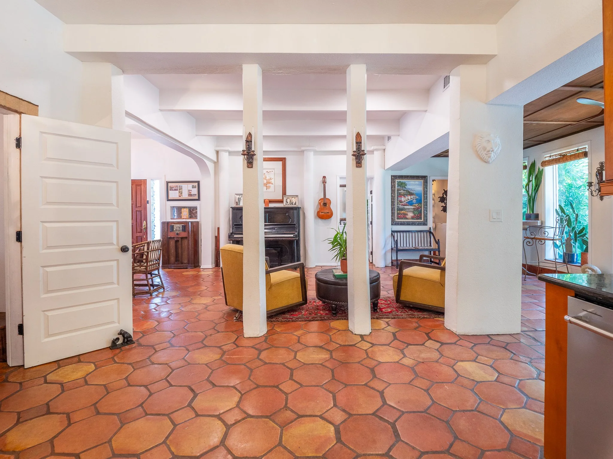 Interior view of a living room with white walls, terracotta tiled floor, a piano, wooden chairs, framed artwork, guitars, plants, and decor.
