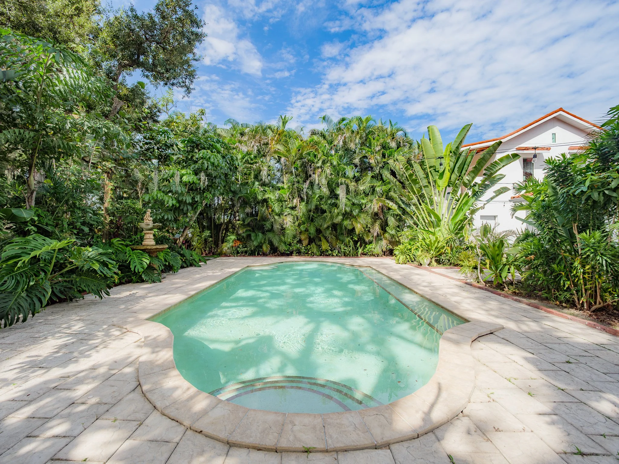 A backyard swimming pool surrounded by a paved deck area, lush tropical plants, trees, and a house with a red-tiled roof in the background. The sky is partly cloudy with blue and white clouds.