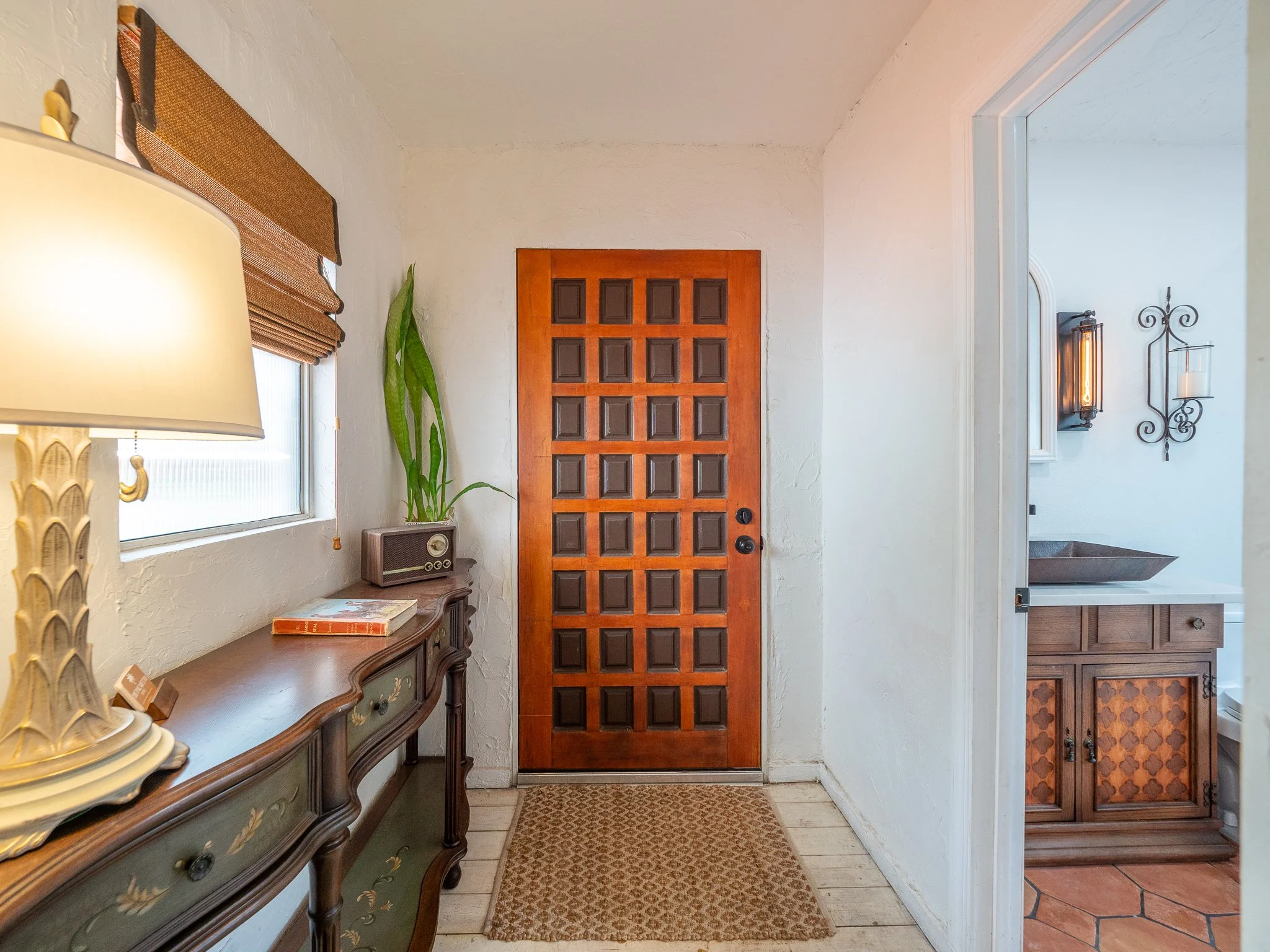 Entryway with wooden door, side table with decorative items, window with blinds, and wall sconces.