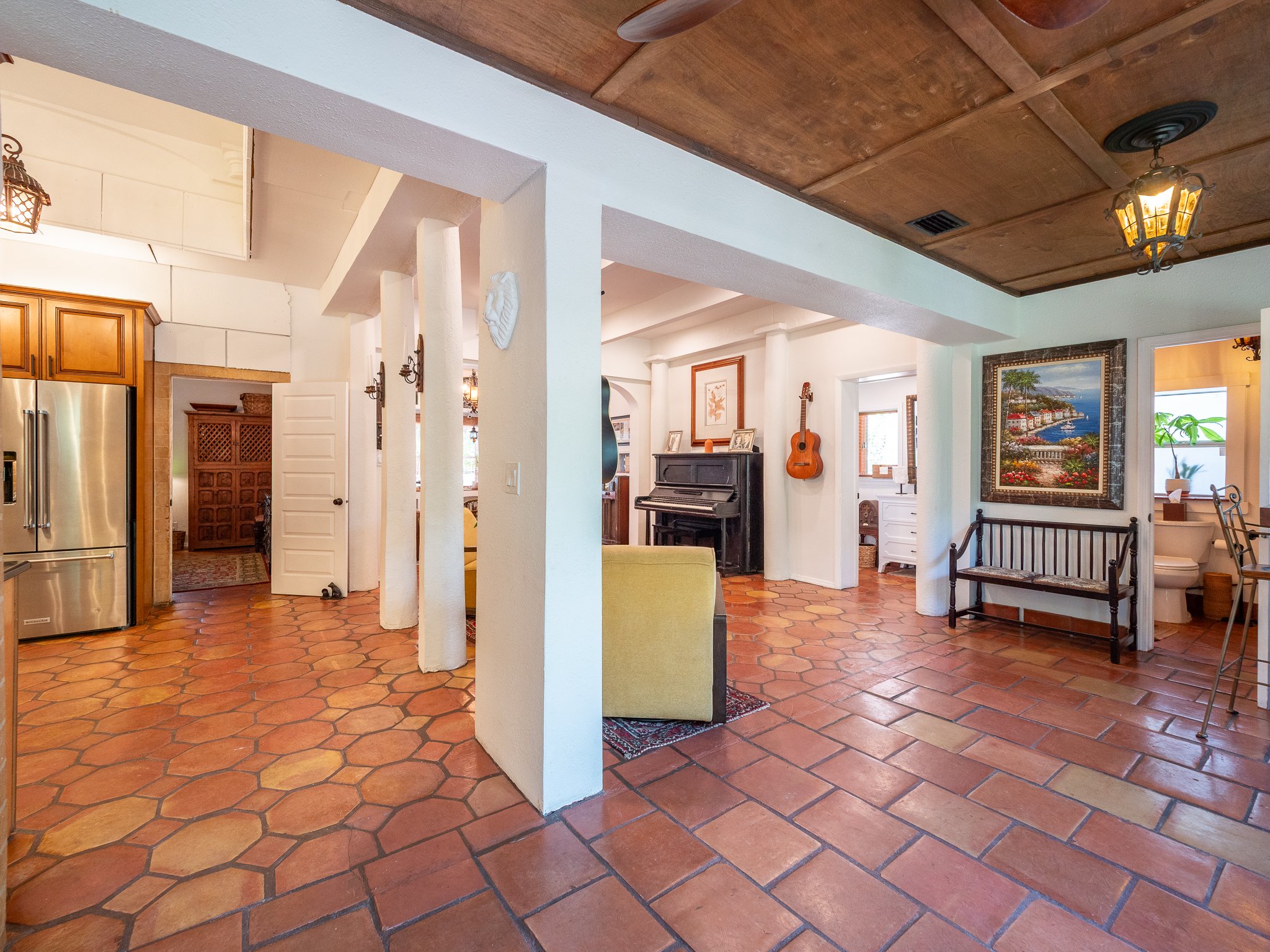 A living room with terra cotta tile floors, white walls, wooden ceiling, and various furniture and decorations including a black piano, a painting of a coastal scene, and a wooden bench.
