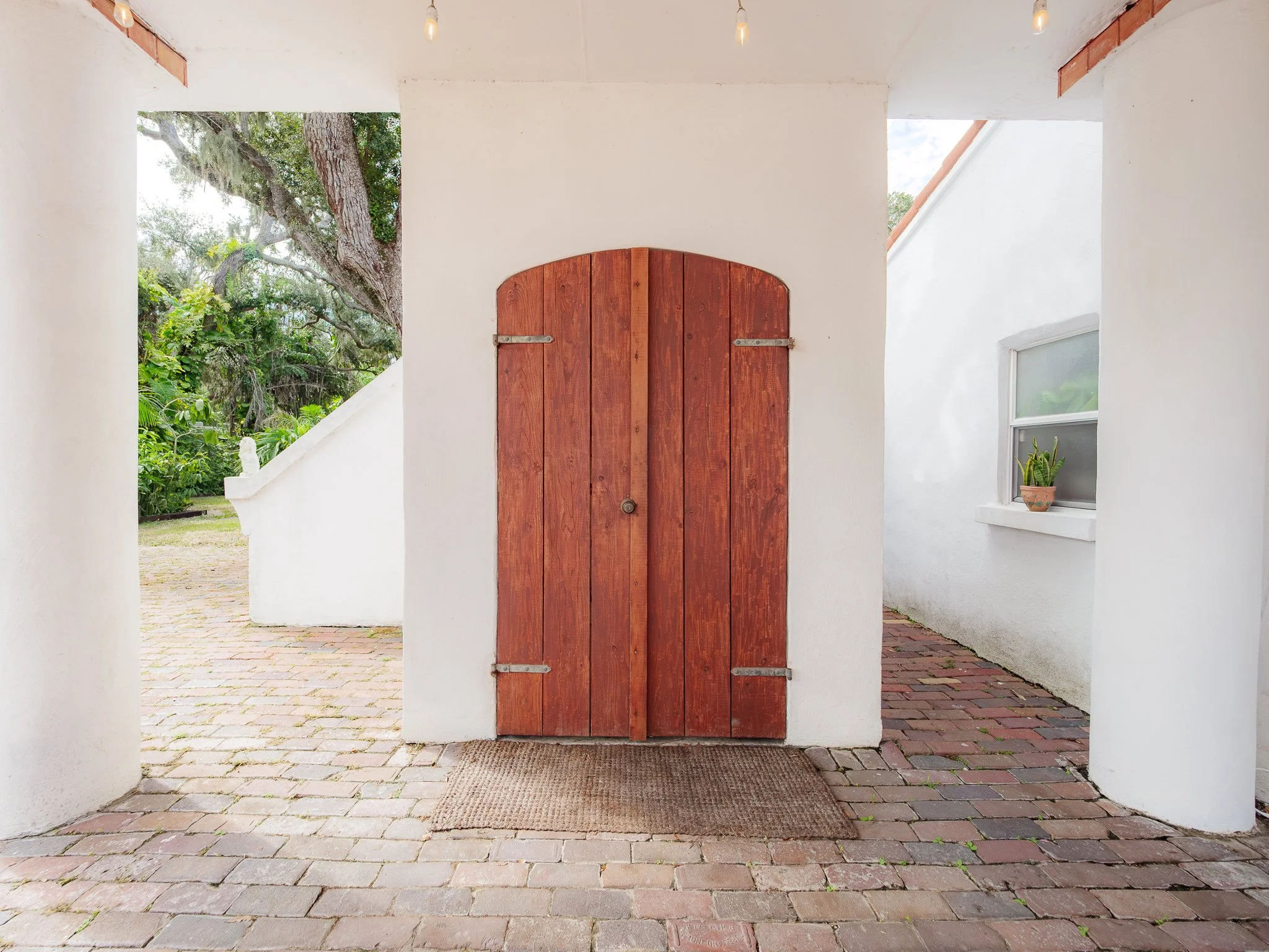 A wooden arched door set in a white stucco wall, with a small mat in front and visible brick paving outside. To the left, there is an outdoor area with trees and greenery.