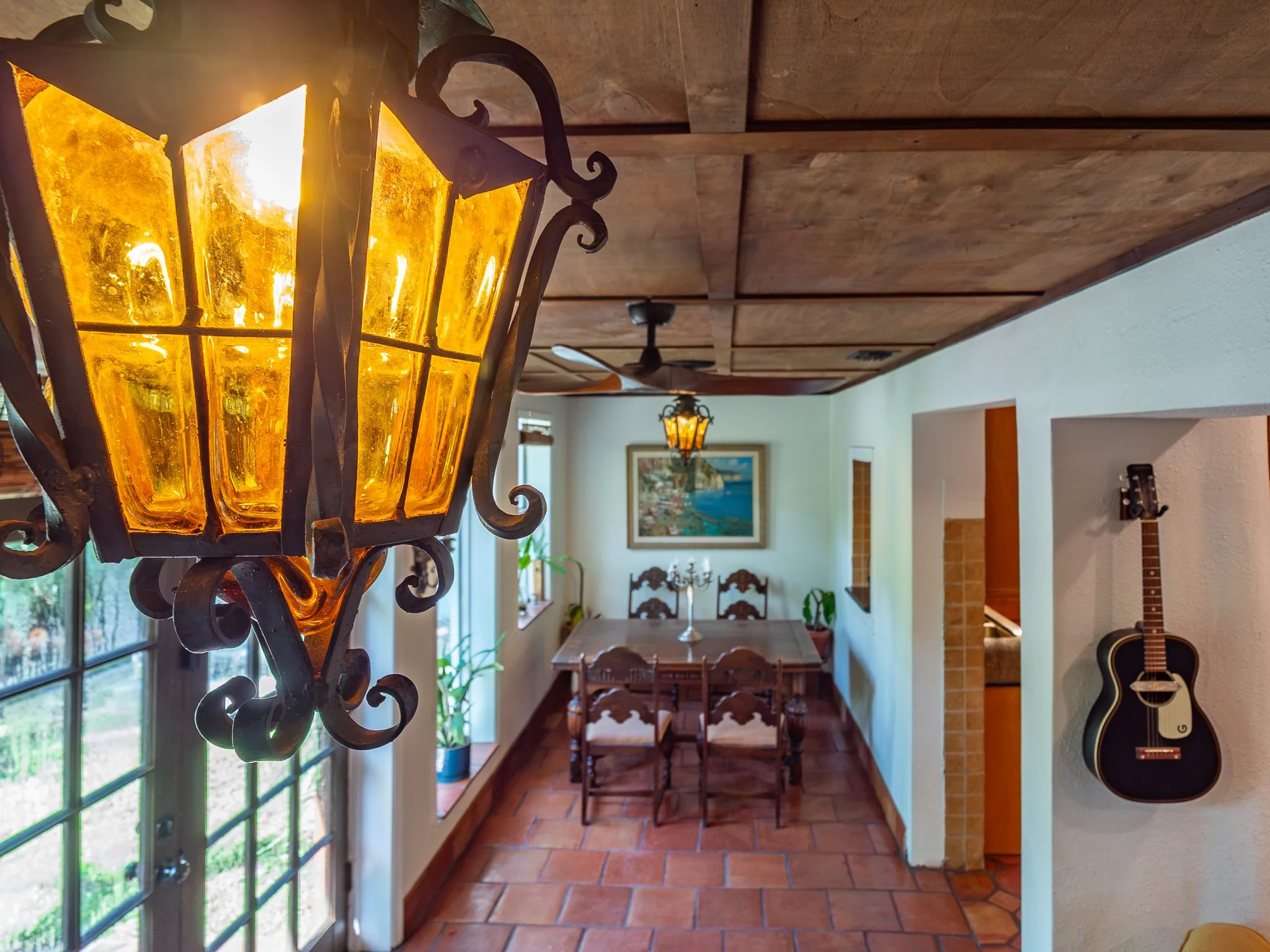 A hanging lantern with yellow-orange glass panels and ornate black metal frame, mounted near a large window. Inside, a ceiling fan with three blades and a chandelier are visible in a dining room with wooden furniture and tiled floors.