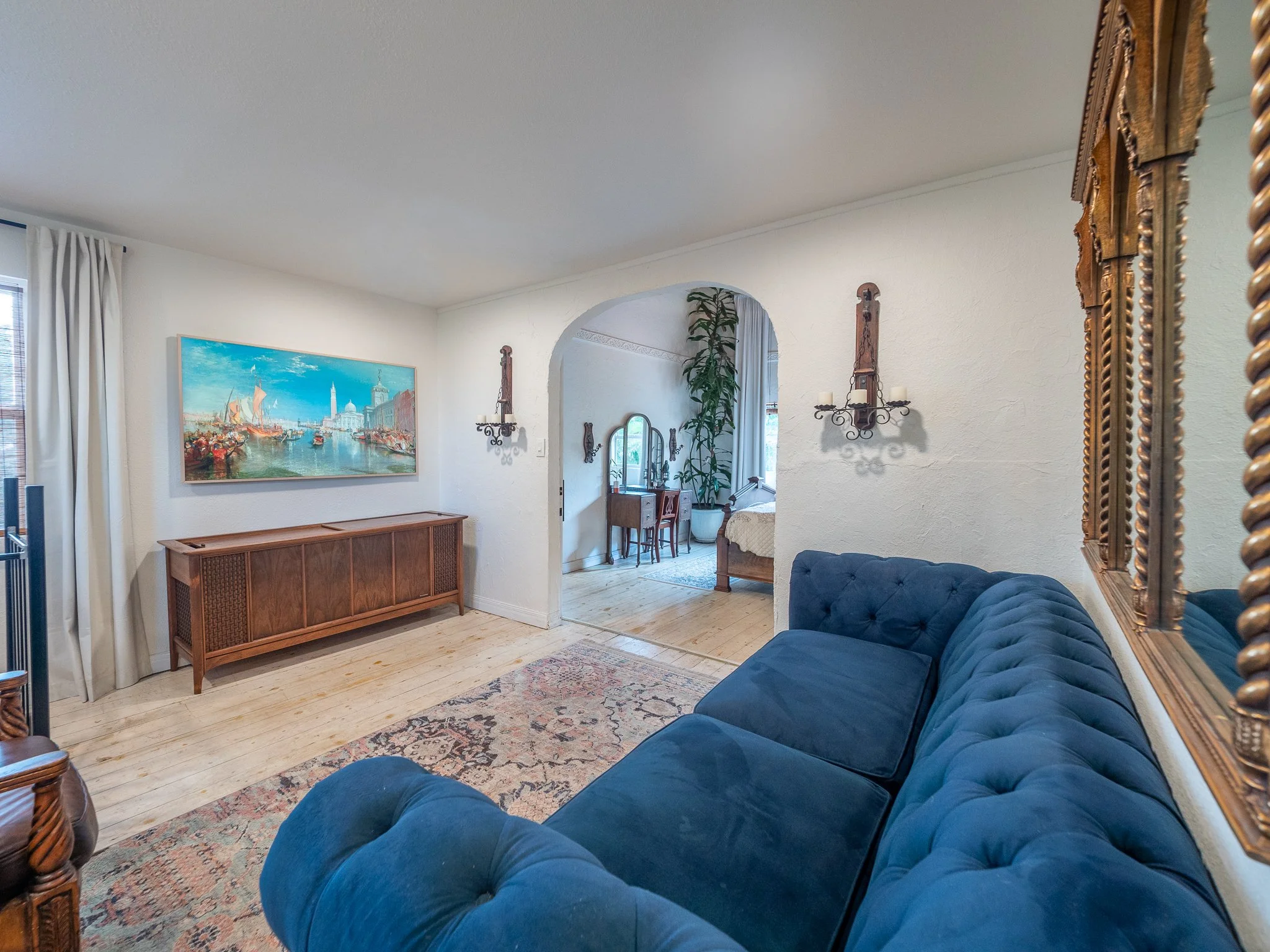 A living room with a large blue tufted sofa in front of a patterned rug, a wooden console under a painting of a harbor scene, and an ornate gold-framed mirror on the right wall, with an arch leading into a dining area.