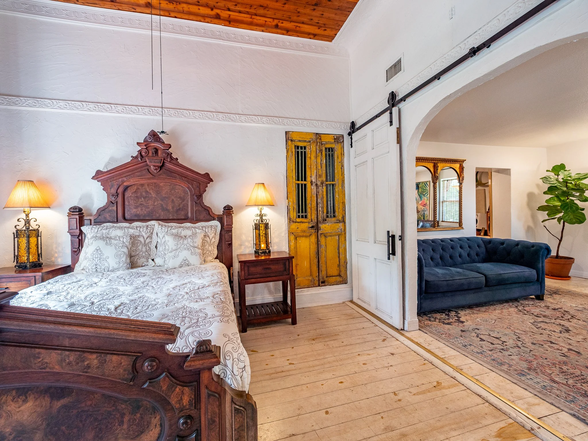 Interior of a living room with a wooden ceiling, white walls, a wooden bed with white patterned bedding, bedside lamps, an antique yellow door, a white sliding door, a blue velvety sofa, a large mirror, a potted plant, and a patterned area rug.