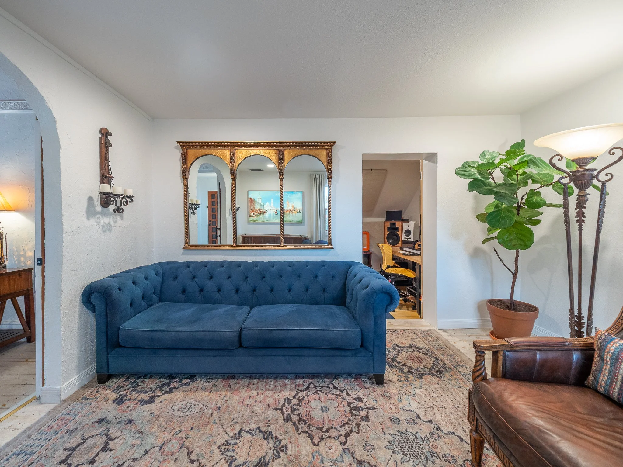 Living room with a blue tufted sofa, a large ornate gold-framed mirror, a plant in a pot, a standing lamp, and a patterned rug.